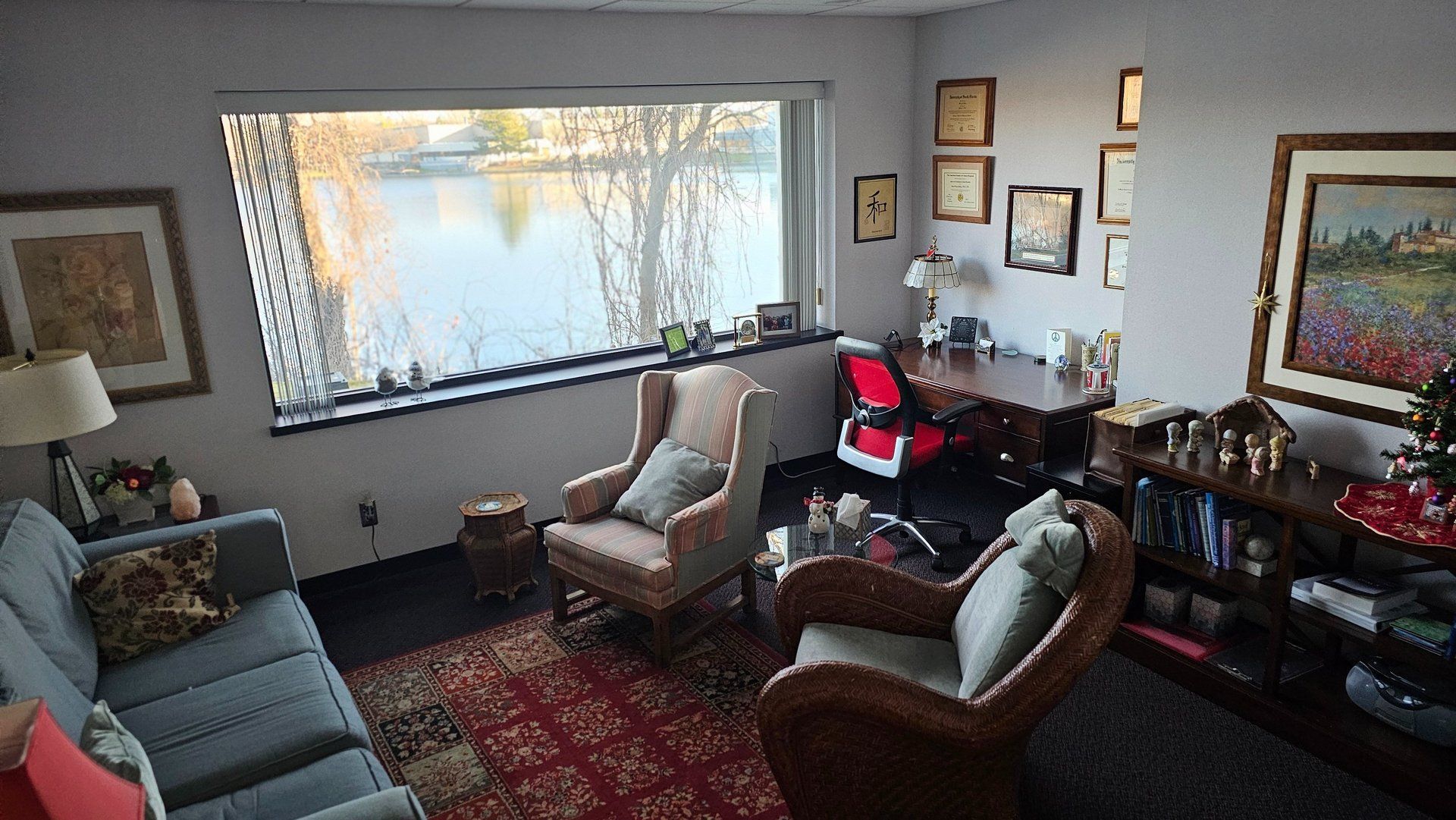 Cozy office with a lake view, featuring seating, desk, and framed art. Red rug and sunlight.