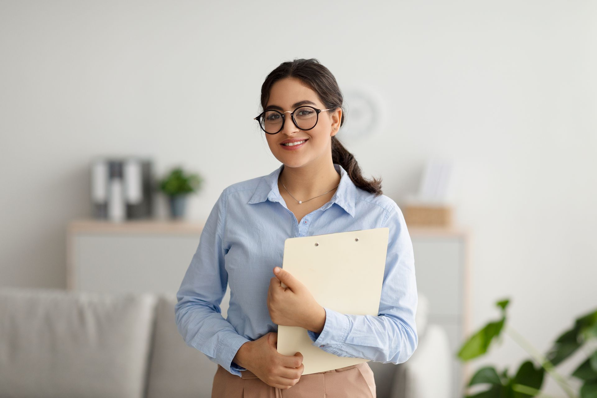 Woman in glasses smiling, holding a folder, in a home office setting.