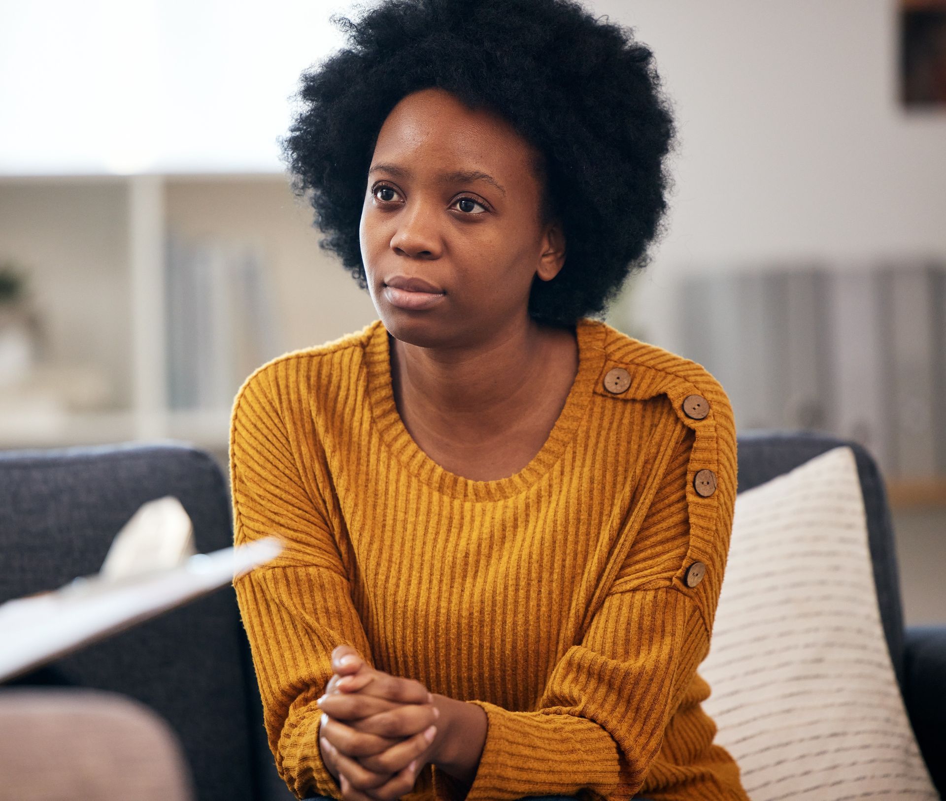 Woman in yellow sweater sits on couch, looking toward a person off-screen.