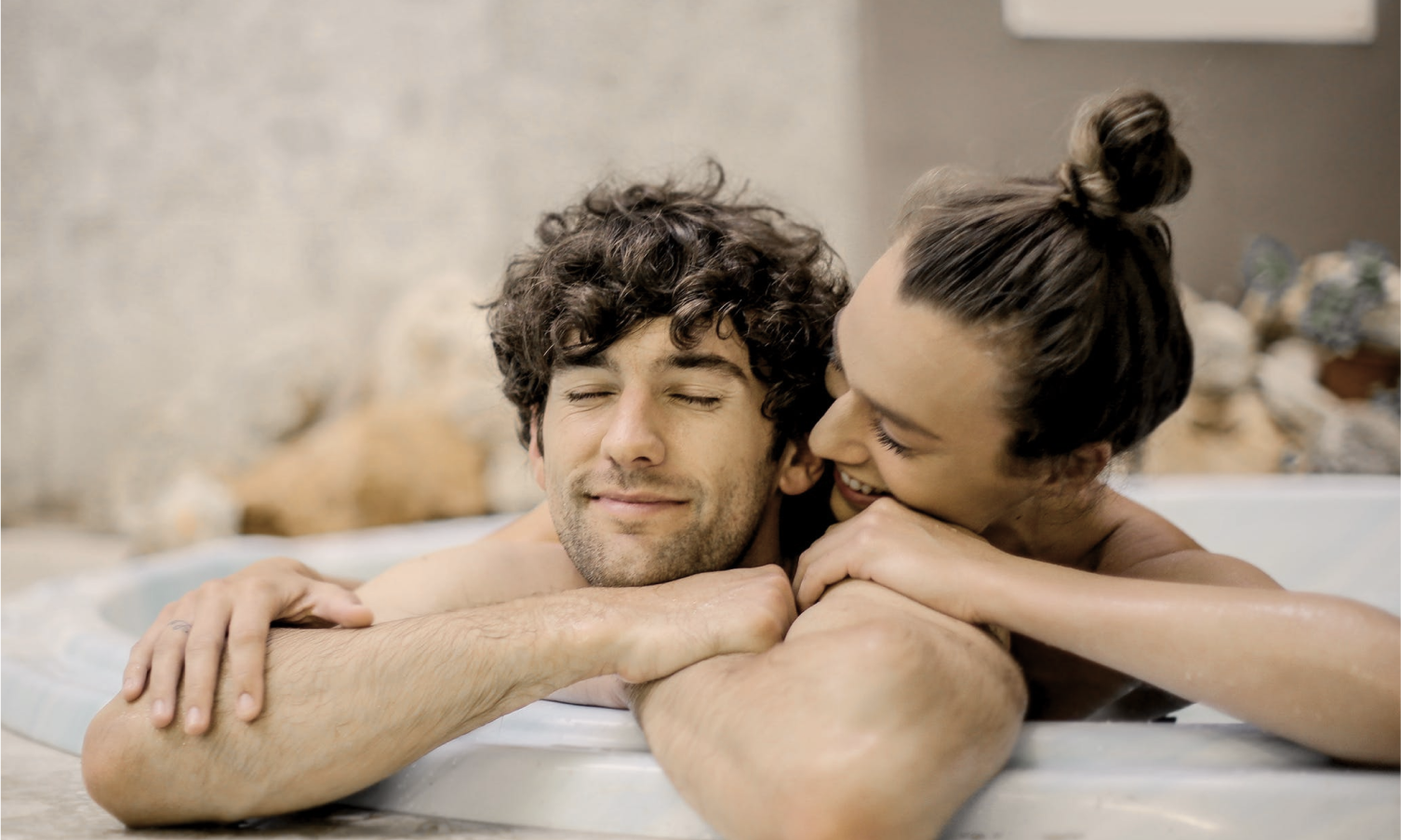 Two people, shoulders up in a jacuzzi, one whispering, the other smiling with eyes closed.