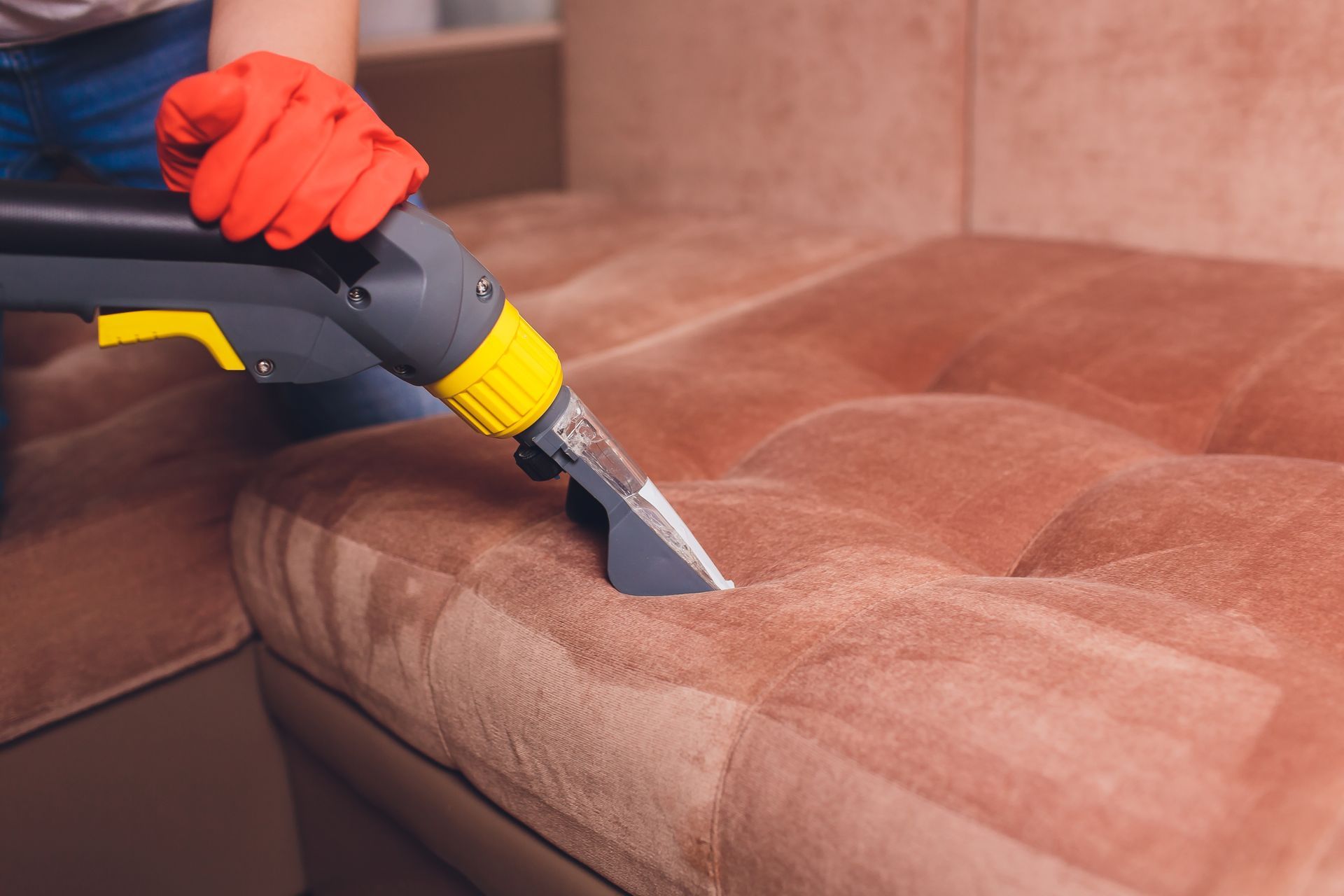 Person in red gloves cleaning brown sofa with an upholstery cleaner.