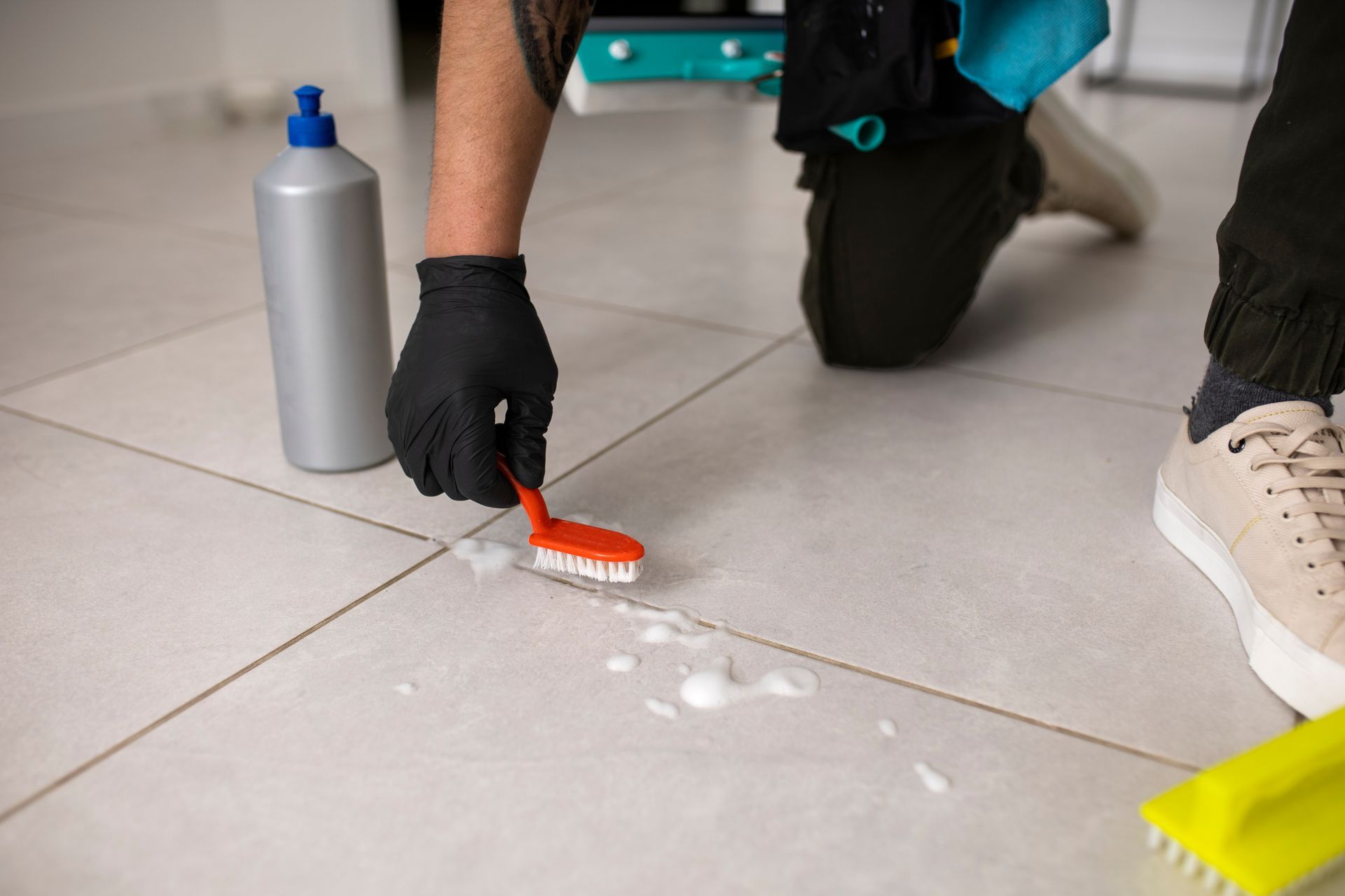 Person kneeling on tile floor scrubbing a white spill with a brush and cleaning supplies nearby
