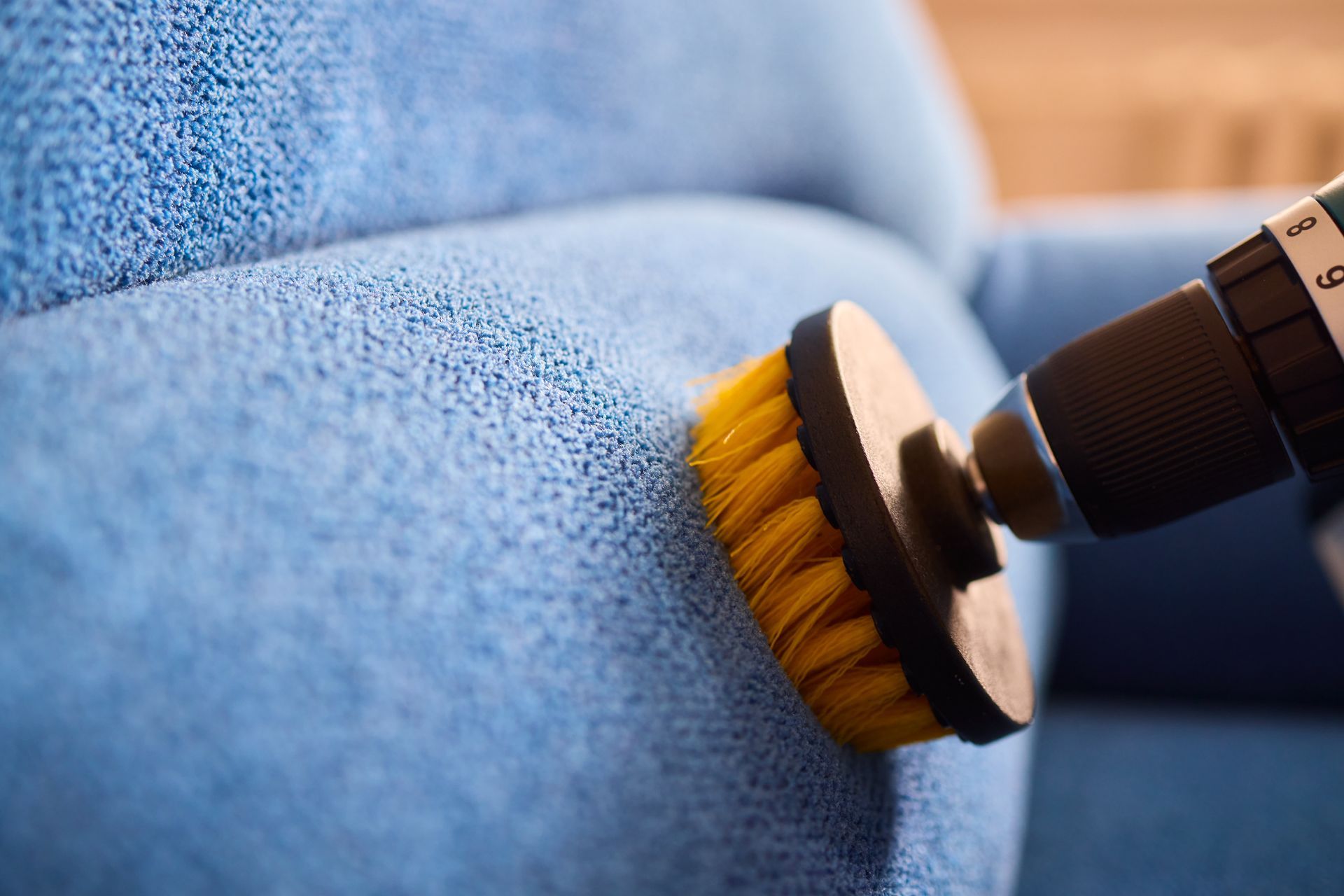 A power drill with brush attachment cleaning a blue upholstered sofa.