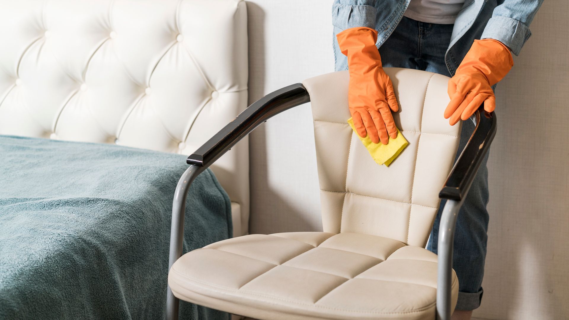 Person in orange gloves cleaning a beige chair with a yellow sponge in a bright bedroom