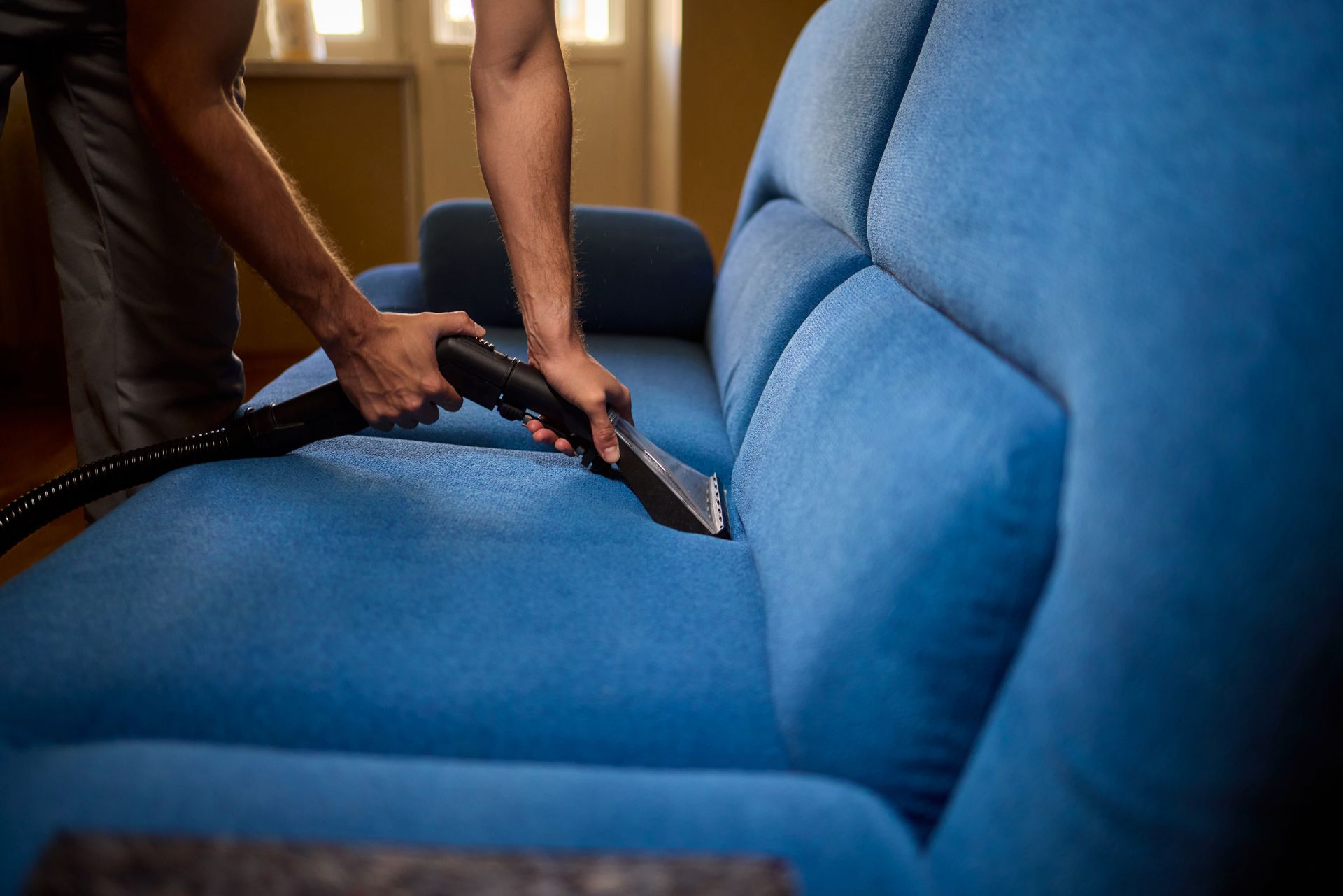 A man is cleaning a blue couch with a vacuum cleaner.