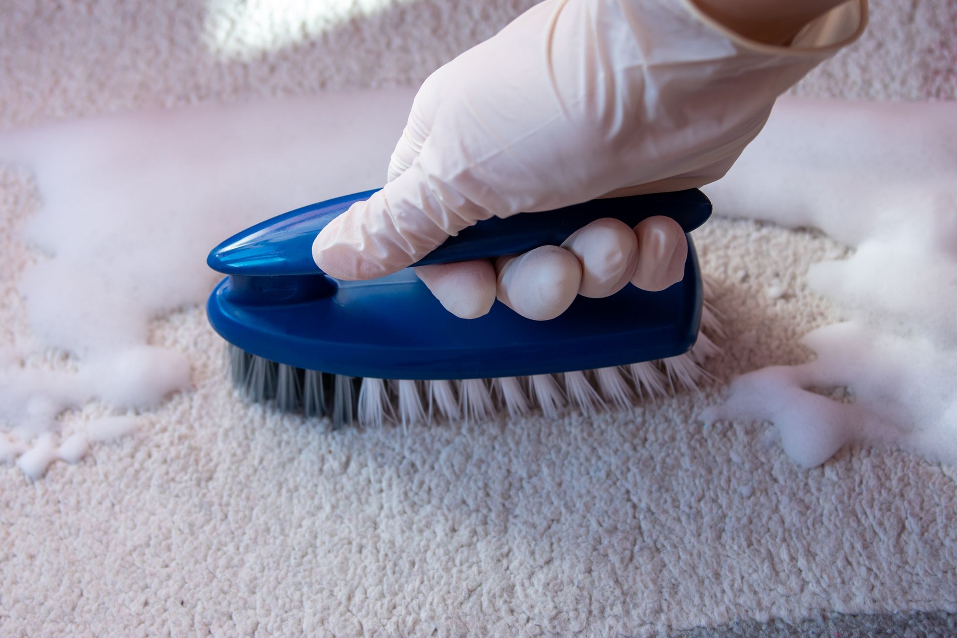 Gloved hand scrubbing a carpet with a blue brush surrounded by white foam.