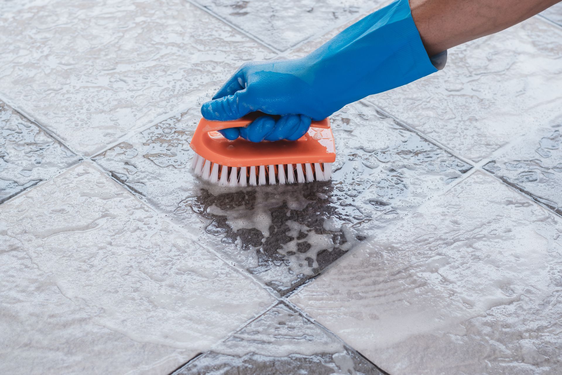 Hand in blue glove scrubs floor tiles with orange brush.