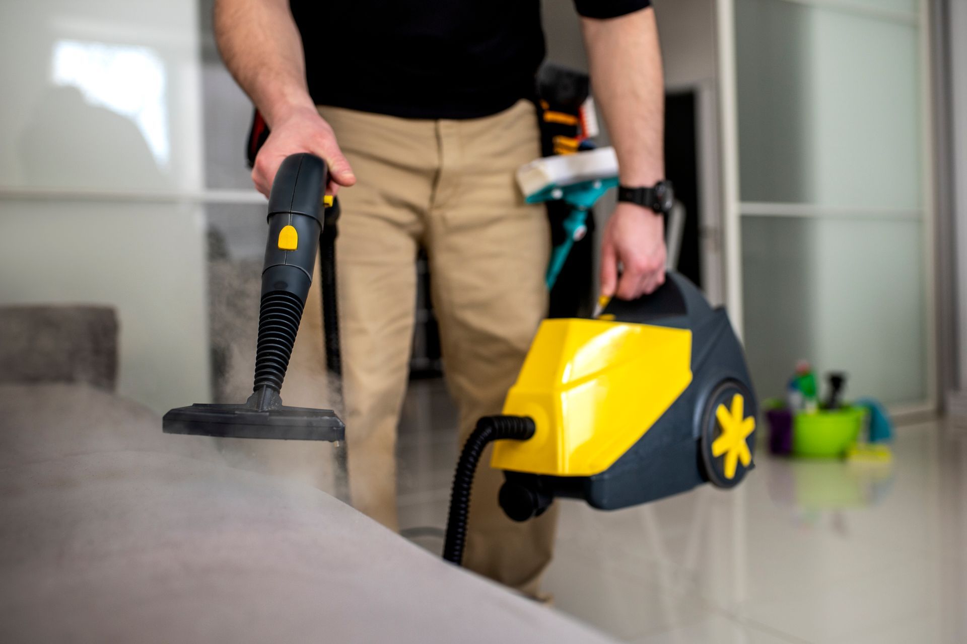 Person steam-cleaning a gray couch with a yellow and black steamer in an apartment.