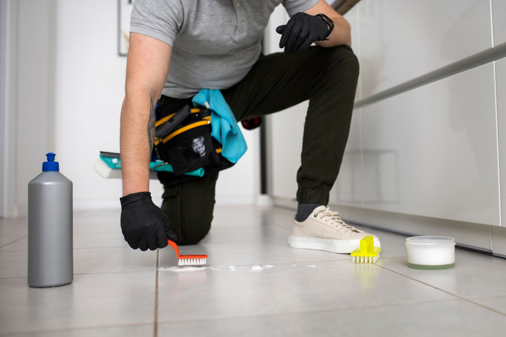 Person cleaning tile floor with brush, cleaning supplies nearby.