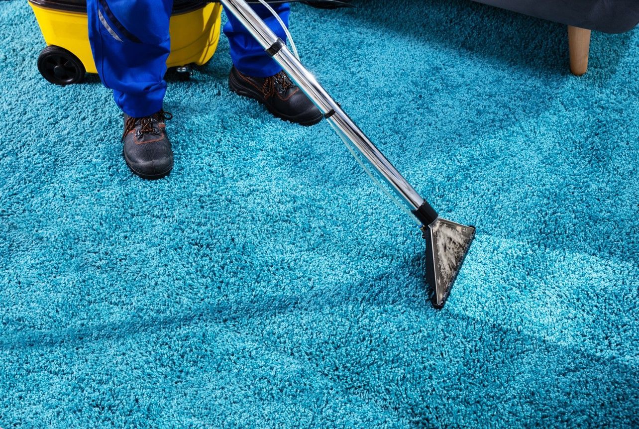 Person in blue cleans a teal carpet with a vacuum cleaner.