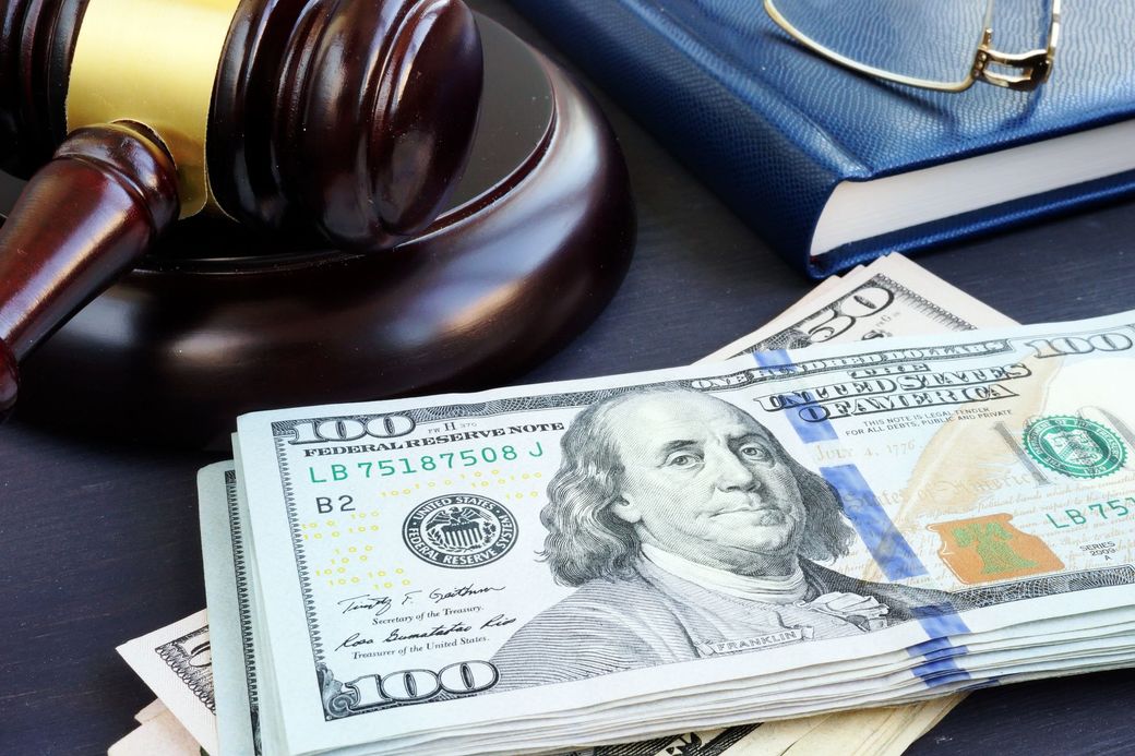 A wooden judge's gavel resting beside a stack of US one-hundred-dollar bills and a blue notebook with glasses.