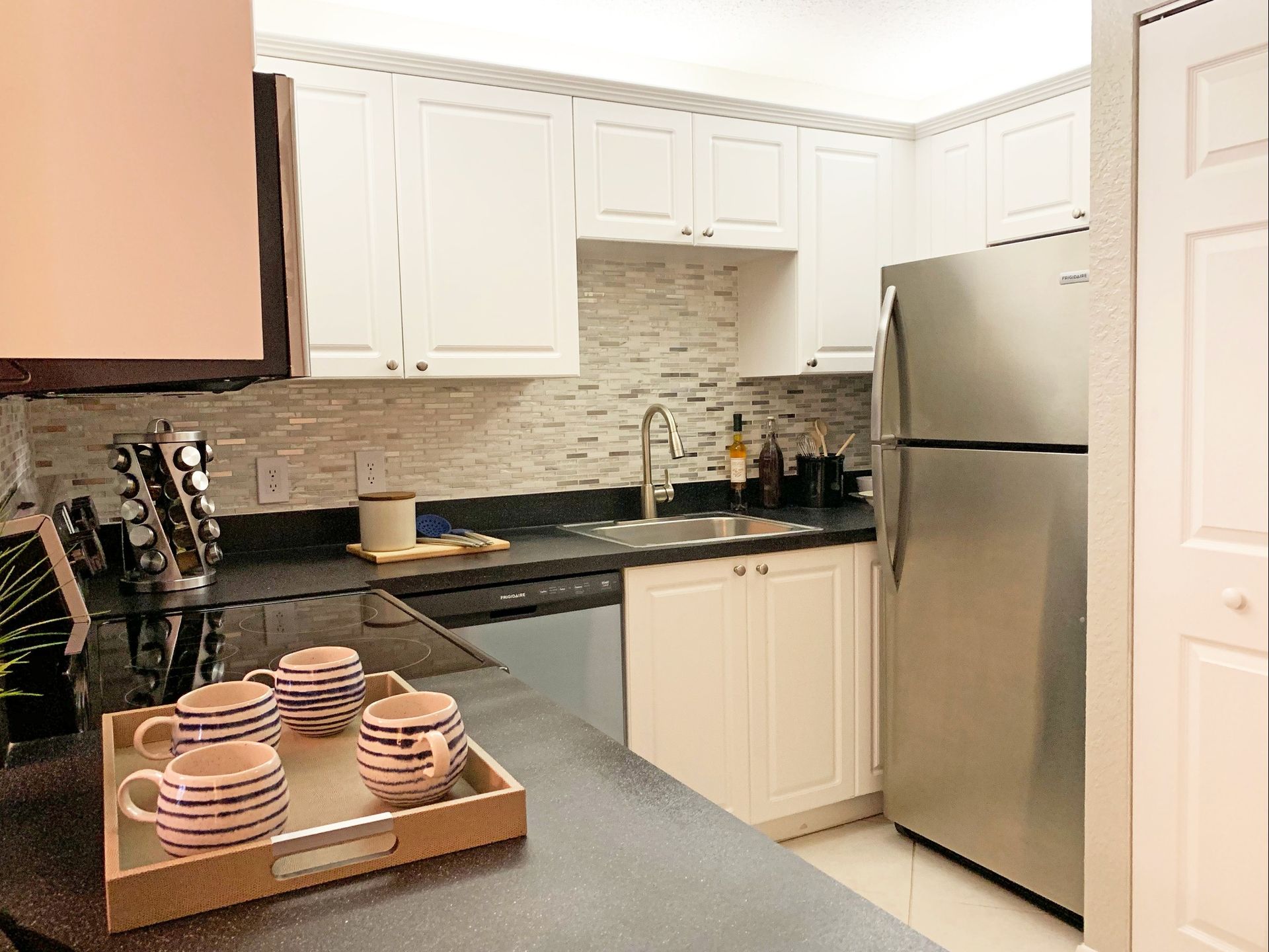 Kitchen with white cabinets, stainless steel refrigerator, and black countertops; a tray of mugs is on the counter.