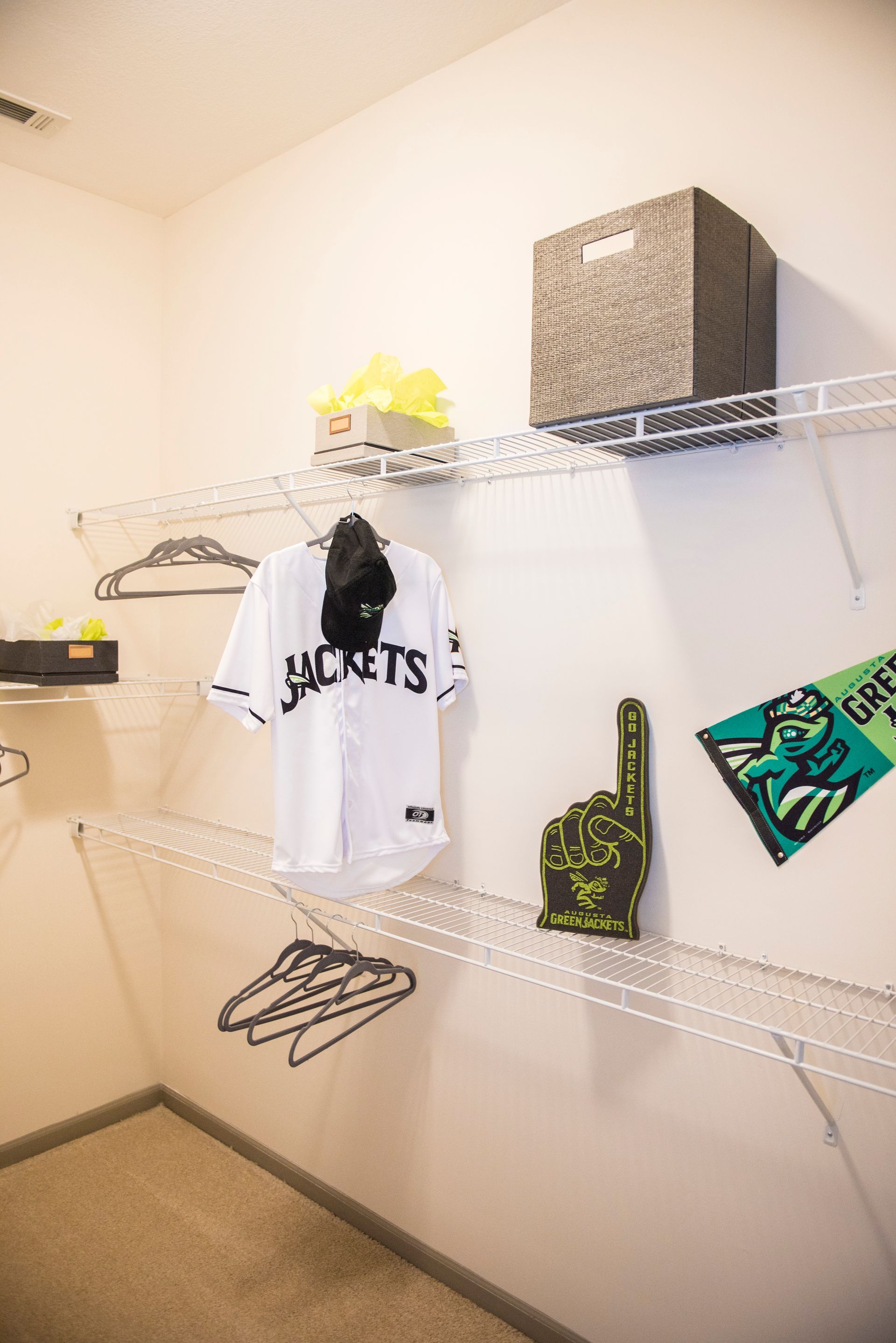 Closet with wire shelving holding a jersey, hat, and fan gear. White walls, beige carpet.