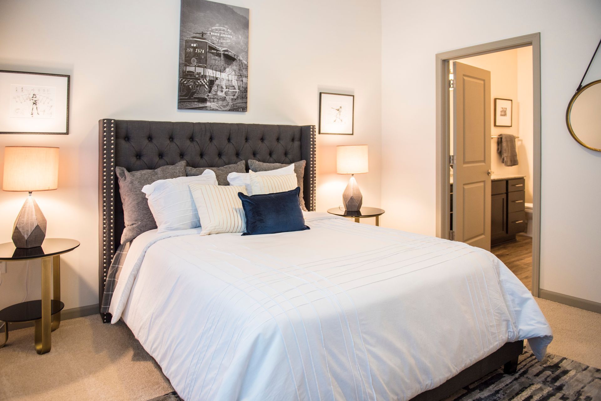 Bedroom with a gray upholstered headboard, lamps on side tables, artwork, and an open bathroom door.