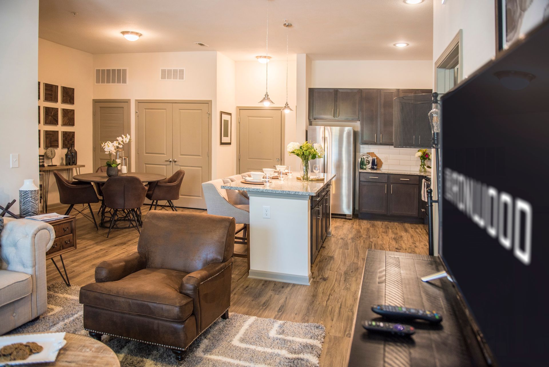 Open-concept apartment interior with living, dining, and kitchen areas. Features include brown leather chair, island, stainless appliances.