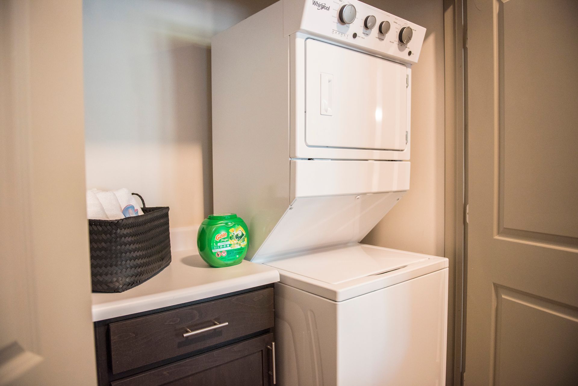 Stacked white washer and dryer in a laundry area, with laundry basket and detergent pod.