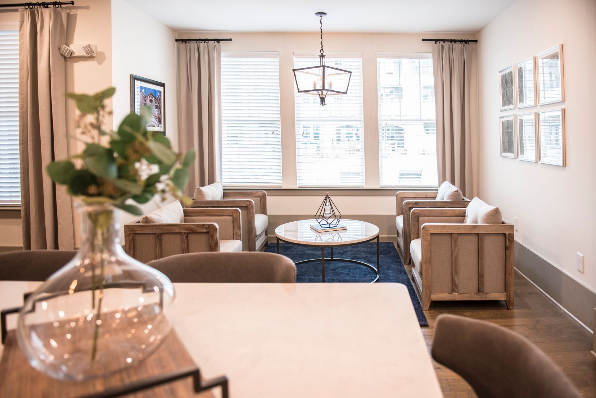 Living room with neutral tones, wood furniture, and a blue rug; a vase of greenery in the foreground.