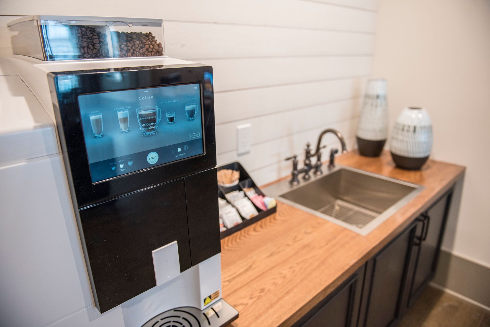 Coffee machine with touch screen on wooden counter, near a sink and decorative vases.