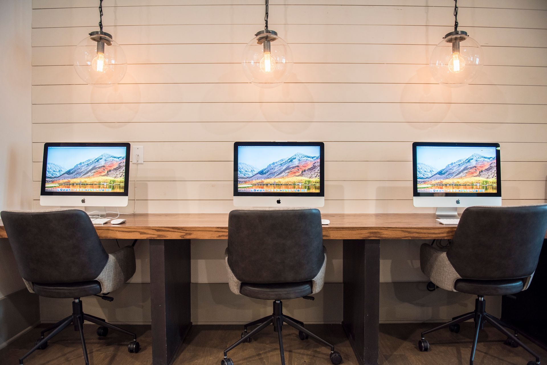 Three desktop computers on a wooden desk in a modern office, with three chairs and pendant lights.