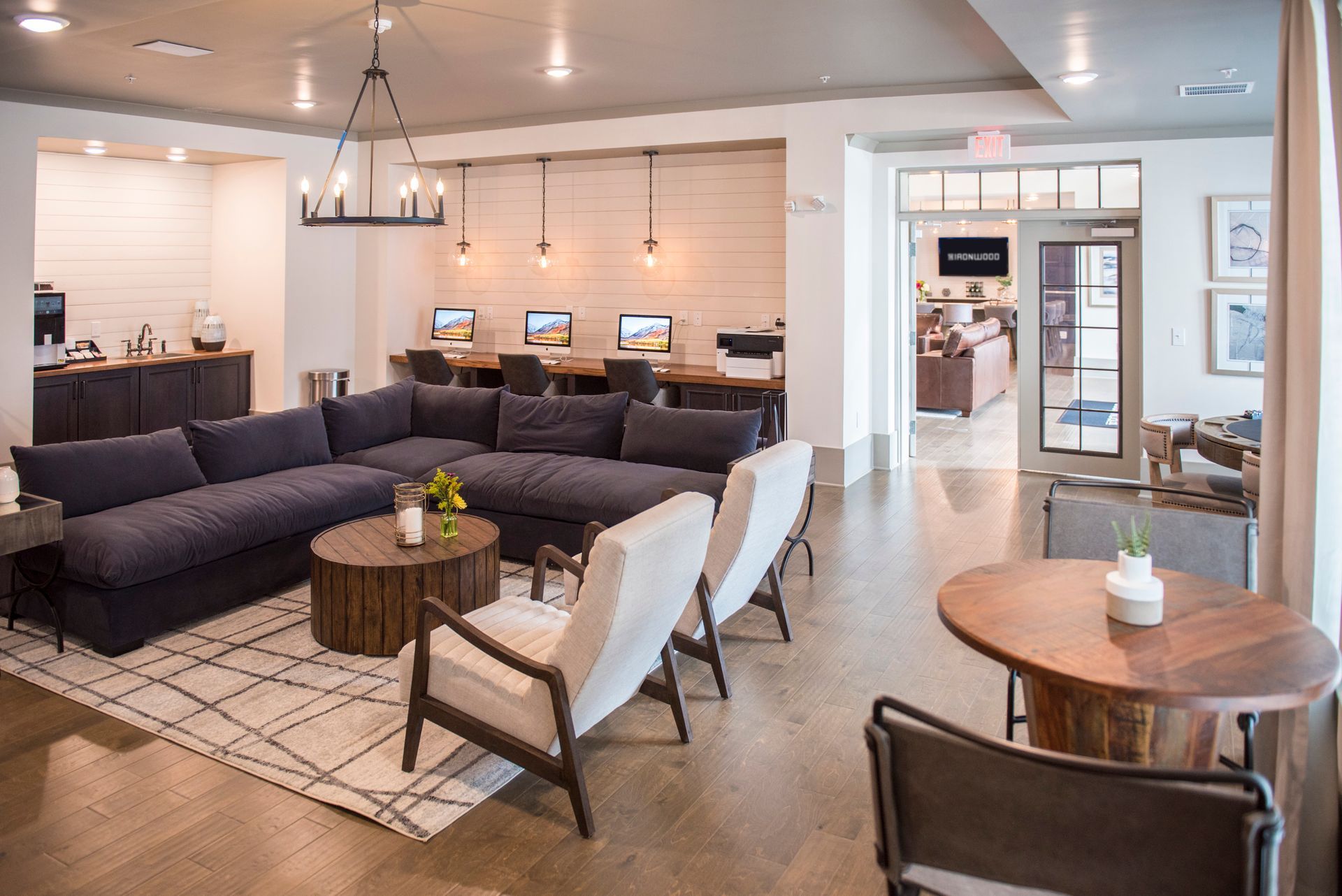 Cozy apartment lounge with sectional sofa, armchairs, computer stations, and small tables; neutral tones.