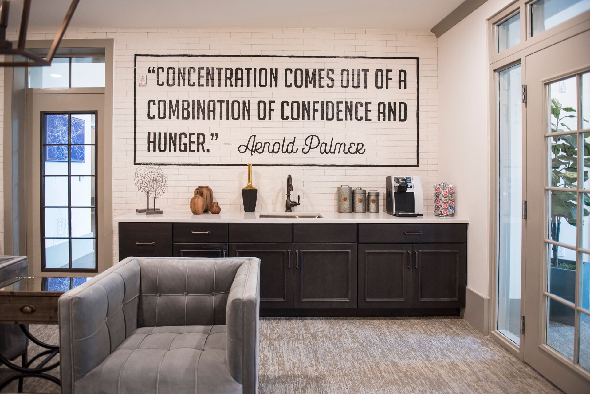A room with a coffee bar, gray armchair, and a sign with a quote by Arnold Palmer.