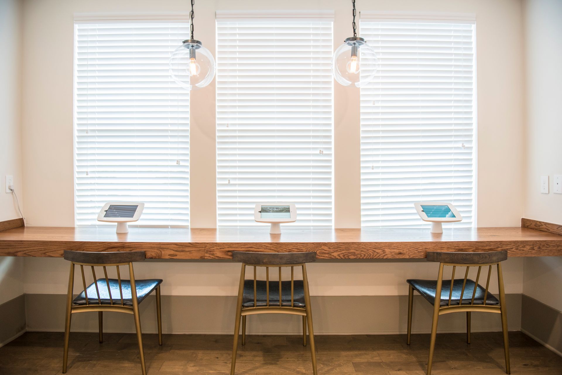 Three tablets on a wood counter with chairs, beneath three windows with blinds.