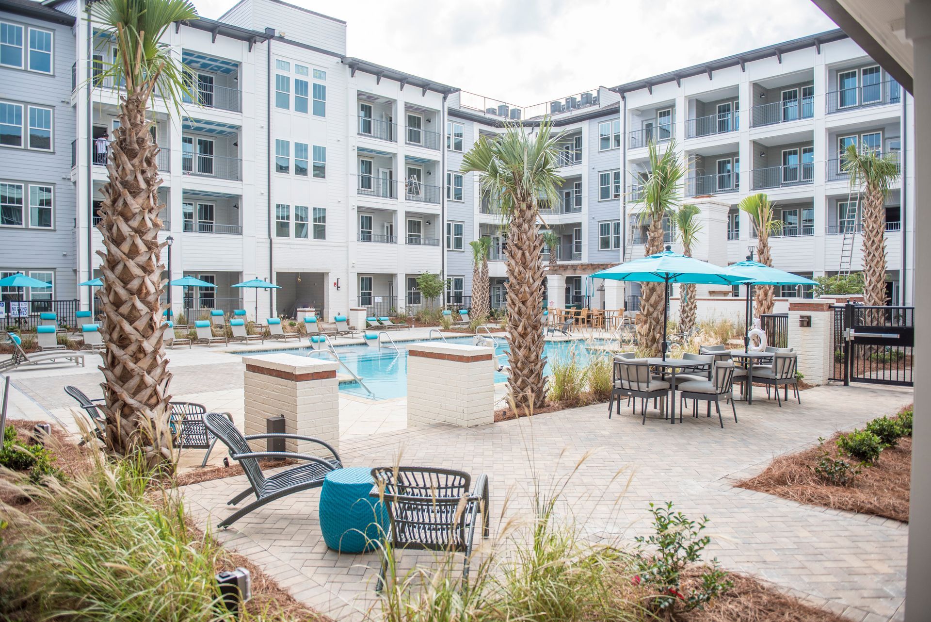 Pool area with lounge chairs, tables, and palm trees, surrounded by white apartment buildings.