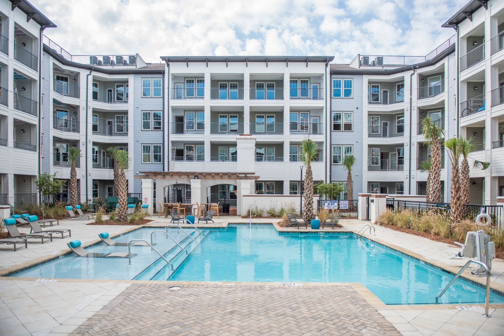Apartment complex with a pool. Buildings are white with balconies. Pool has lounge chairs and palm trees.