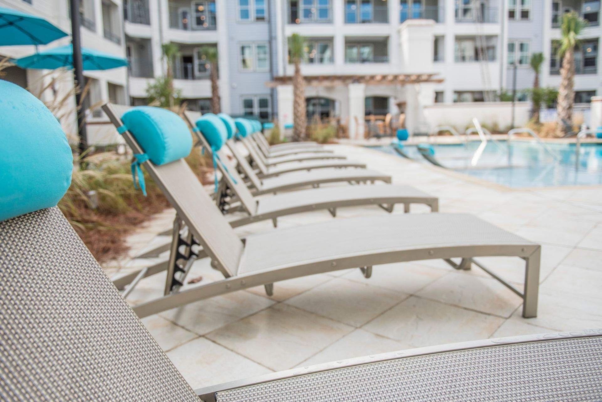 Lounge chairs with turquoise cushions near a pool. Modern apartment buildings in the background.