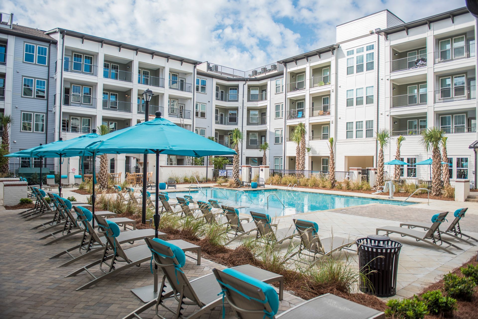 Pool area with lounge chairs, blue umbrellas, and a multi-story apartment building.