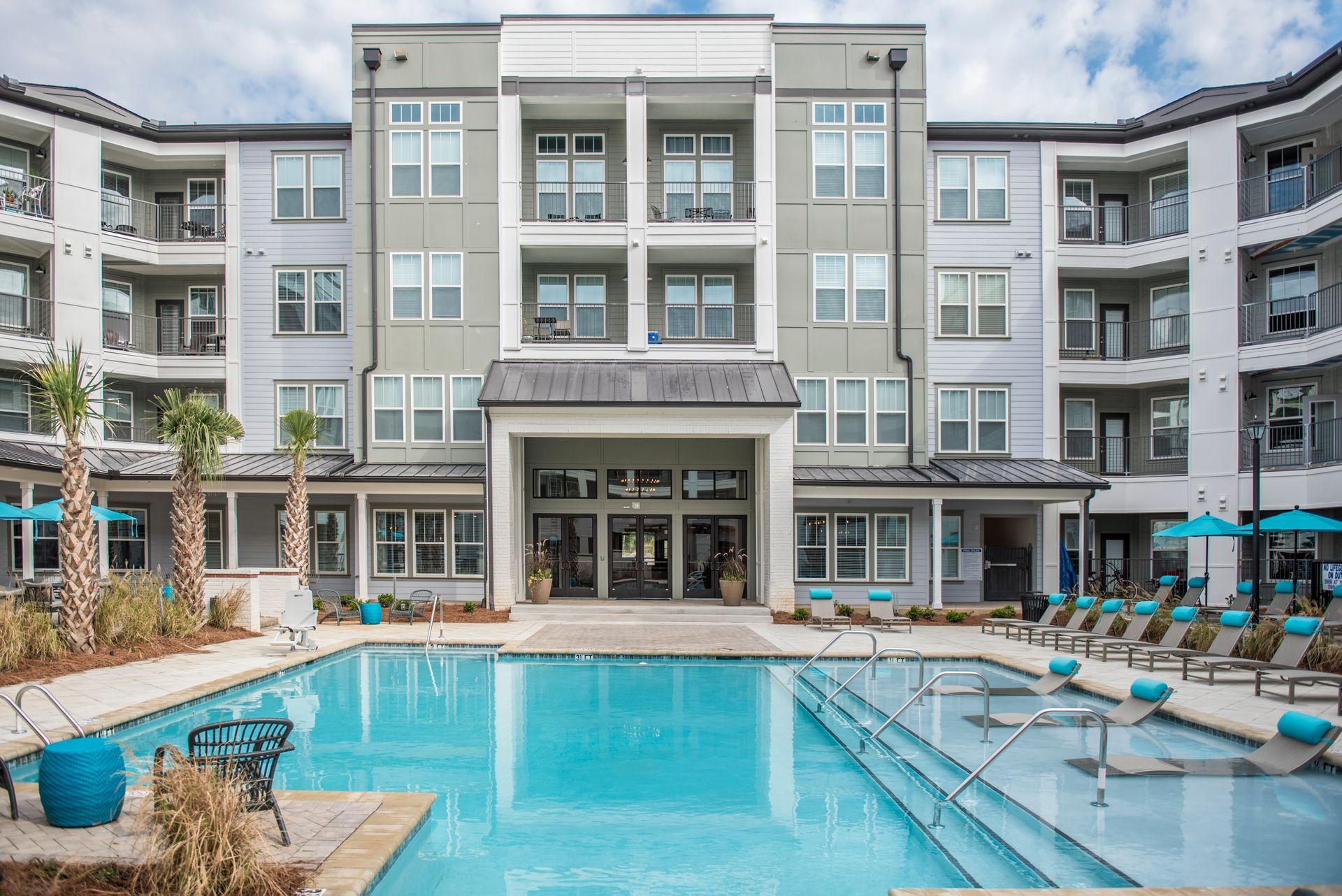 Apartment building with pool. Exterior view: grey building, turquoise pool, lounge chairs, blue umbrellas.