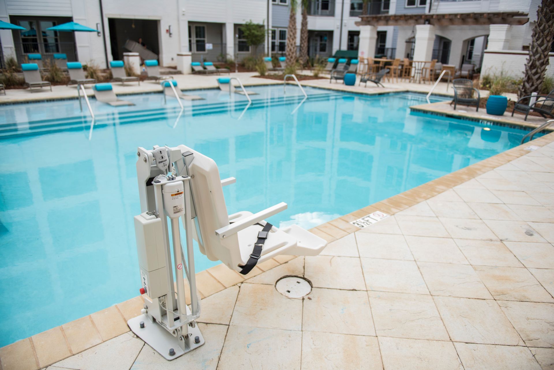 Poolside chair lift; cream-colored chair, blue pool, light stone tiles, apartment building in background.