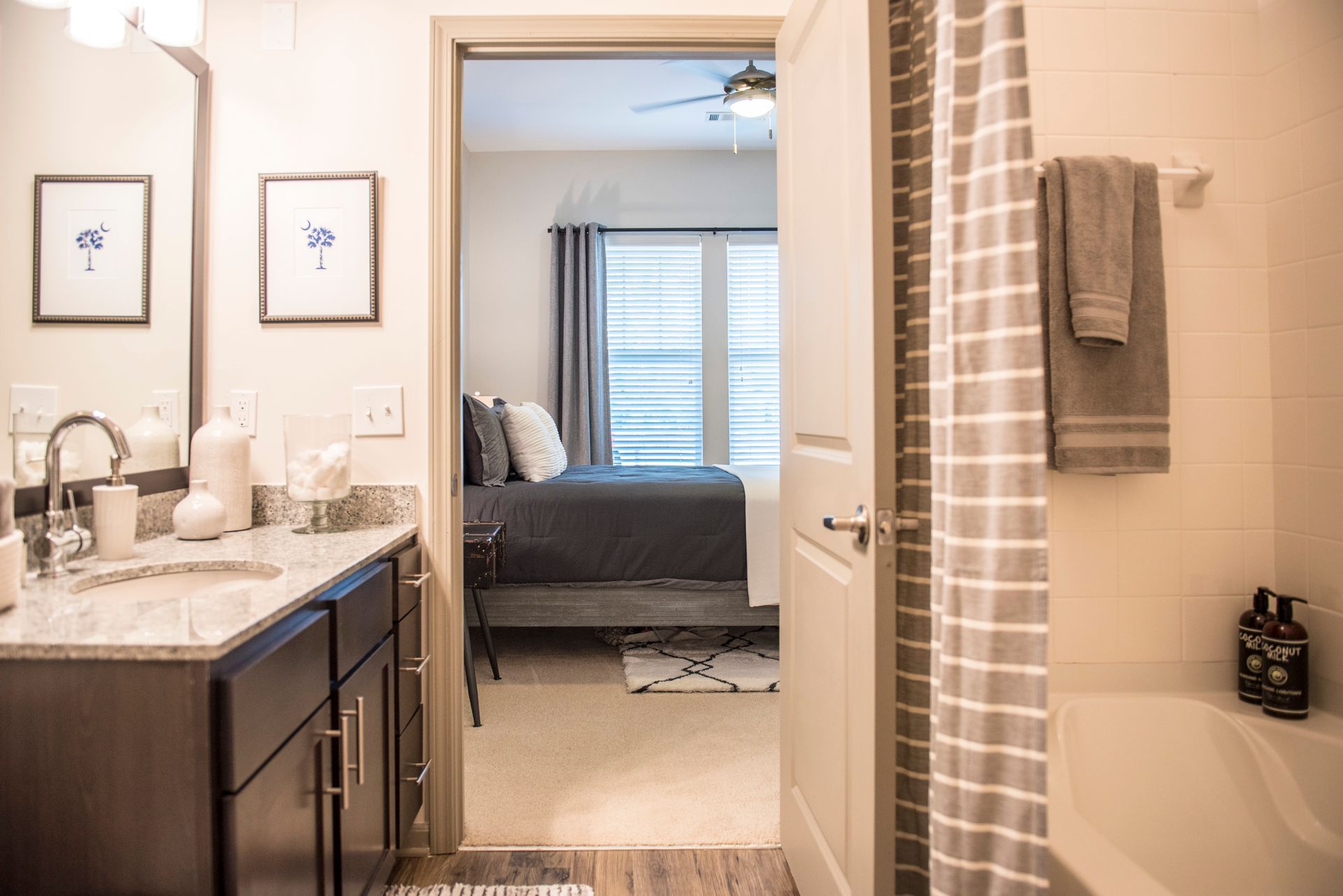 Bathroom doorway leads to bedroom with grey bedding. White walls, grey accents.