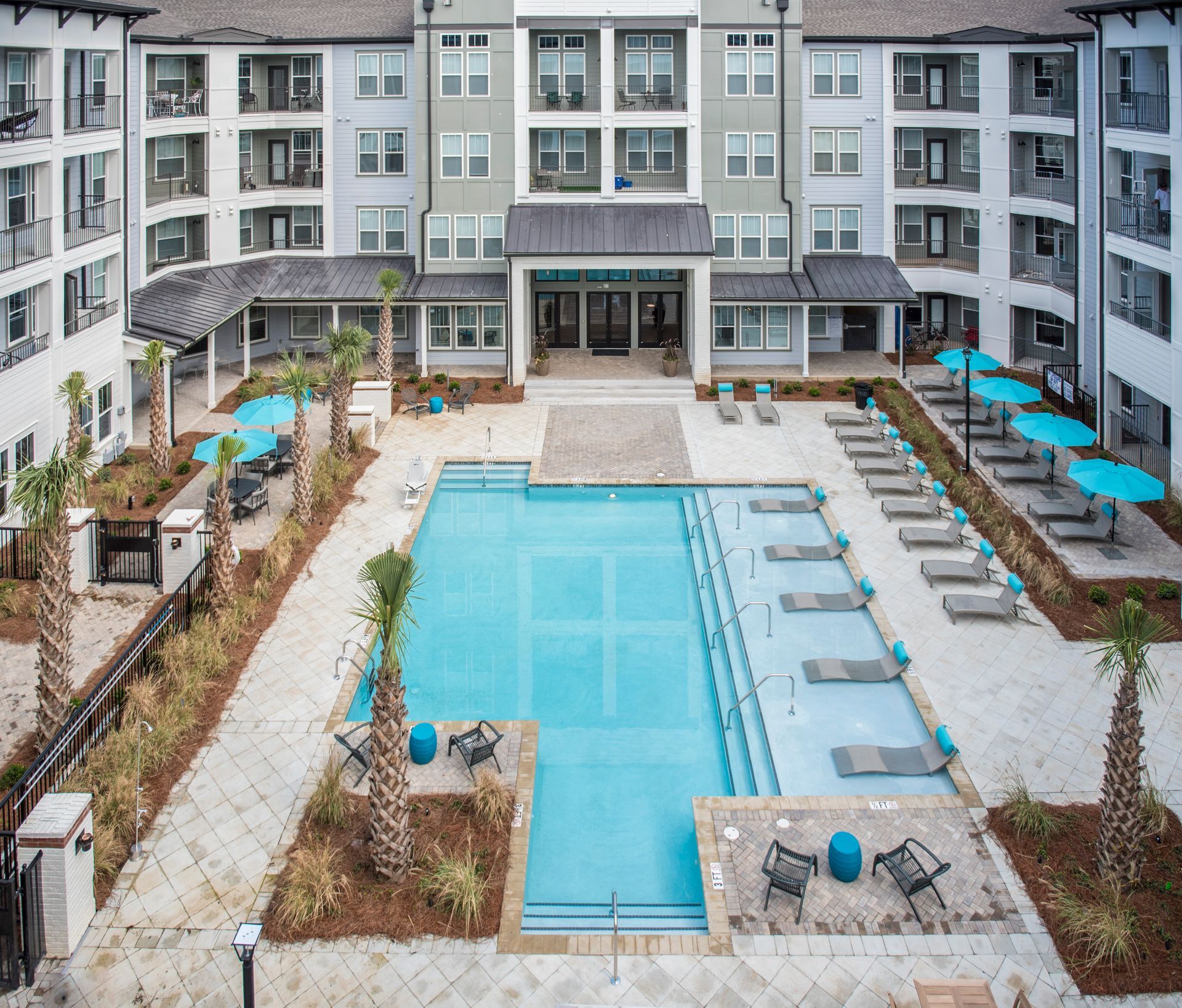 Aerial view of apartment complex with pool; light blue water, lounge chairs, and palm trees.