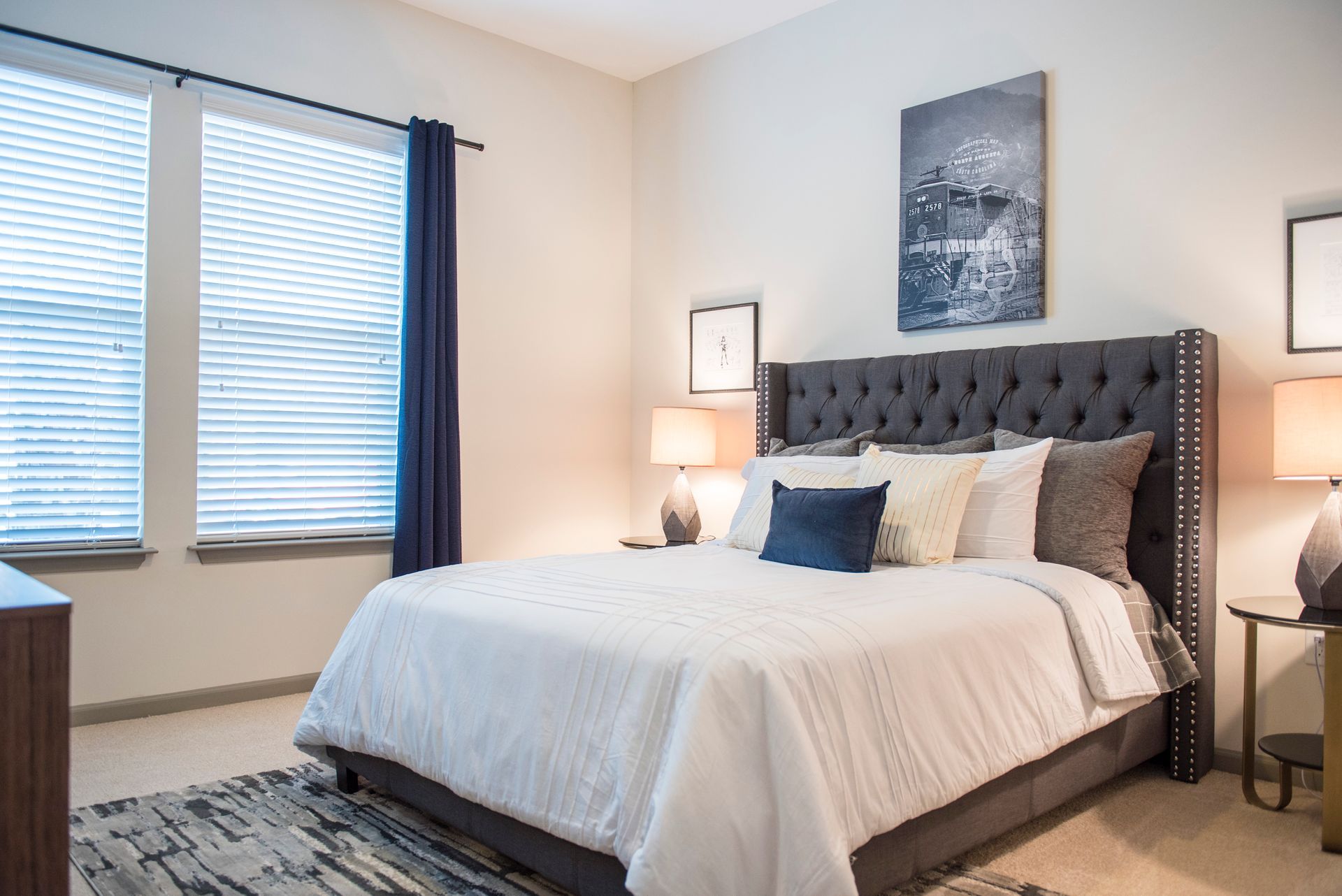 Bedroom with a bed, two windows, art, and bedside tables; beige walls, dark blue accents.