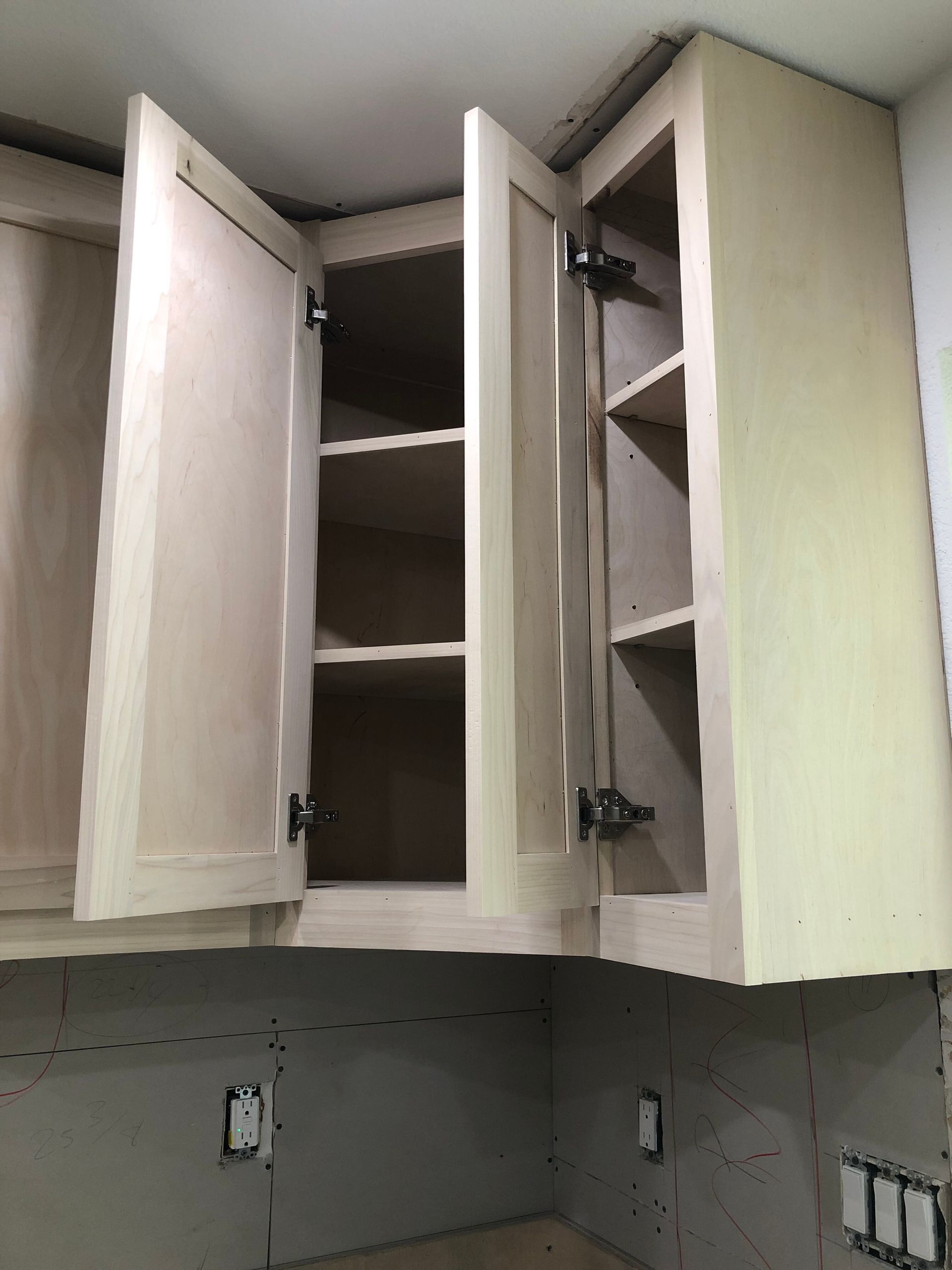 Unfinished light-colored wooden kitchen cabinets installed above a wall with exposed gray drywall and electrical outlets.