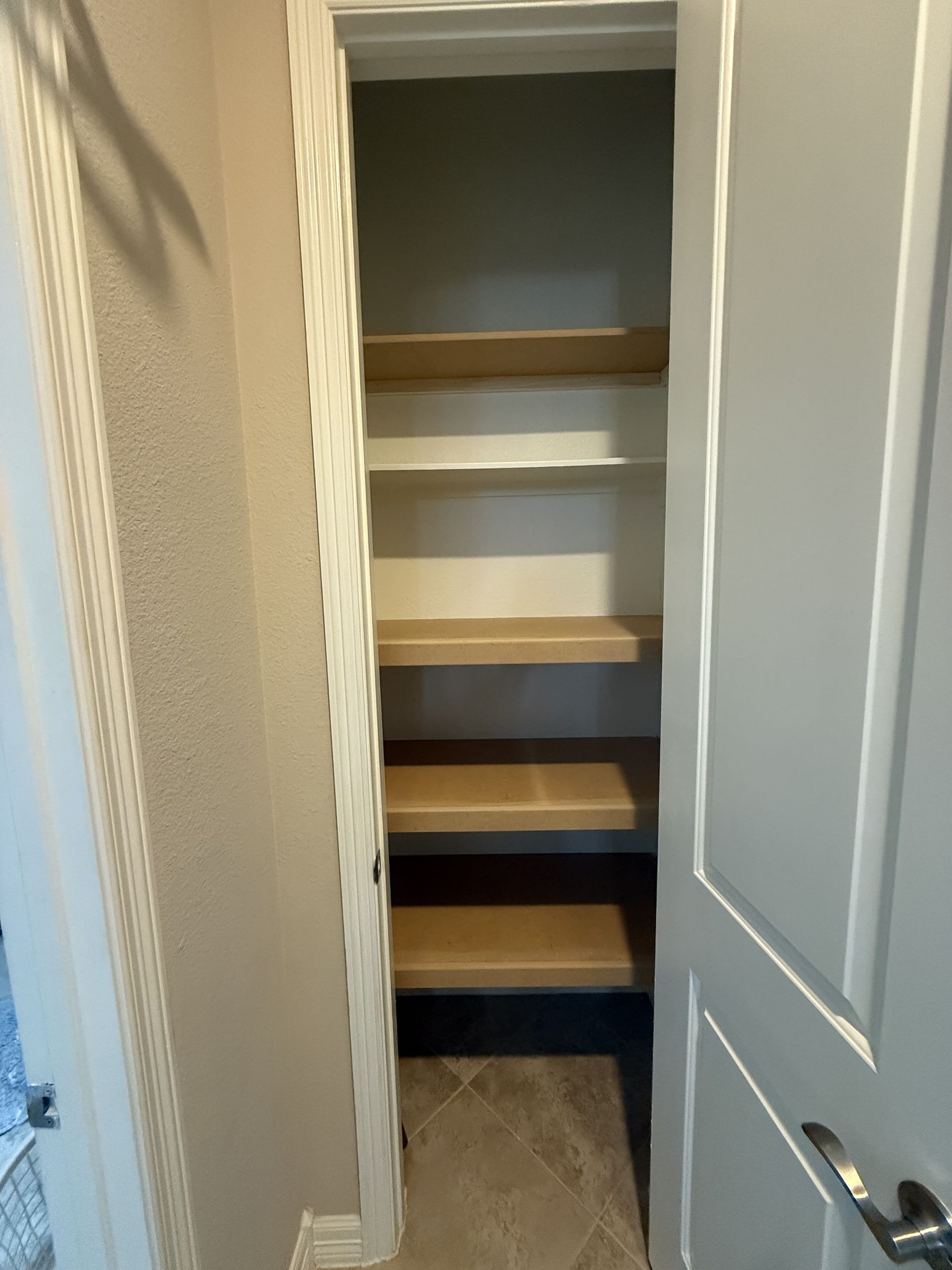 A narrow, open-doored pantry closet featuring light-colored wooden shelves against a light gray wall.