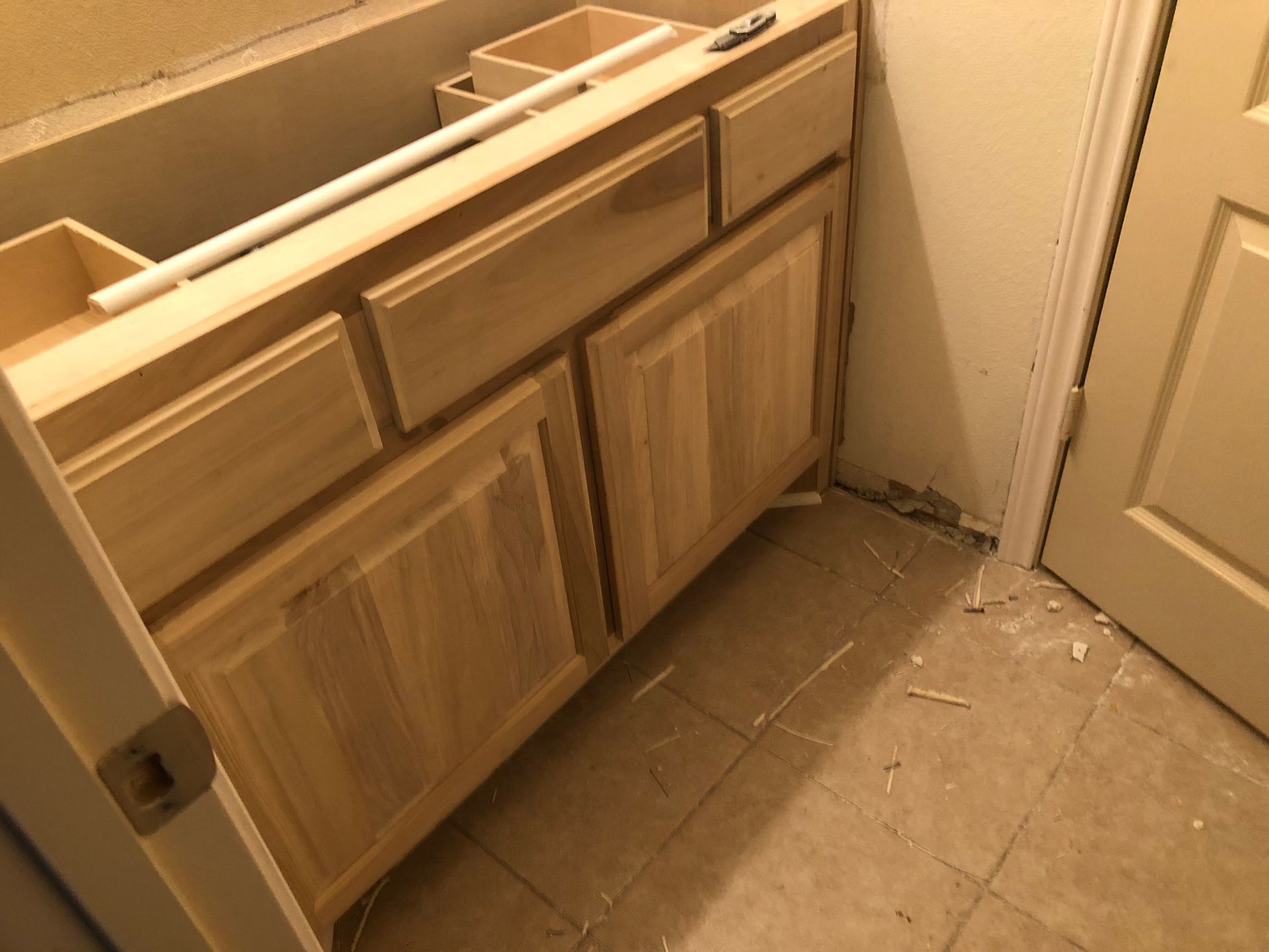 A wooden bathroom vanity cabinet being installed, placed against a wall on a tiled floor near a doorway.