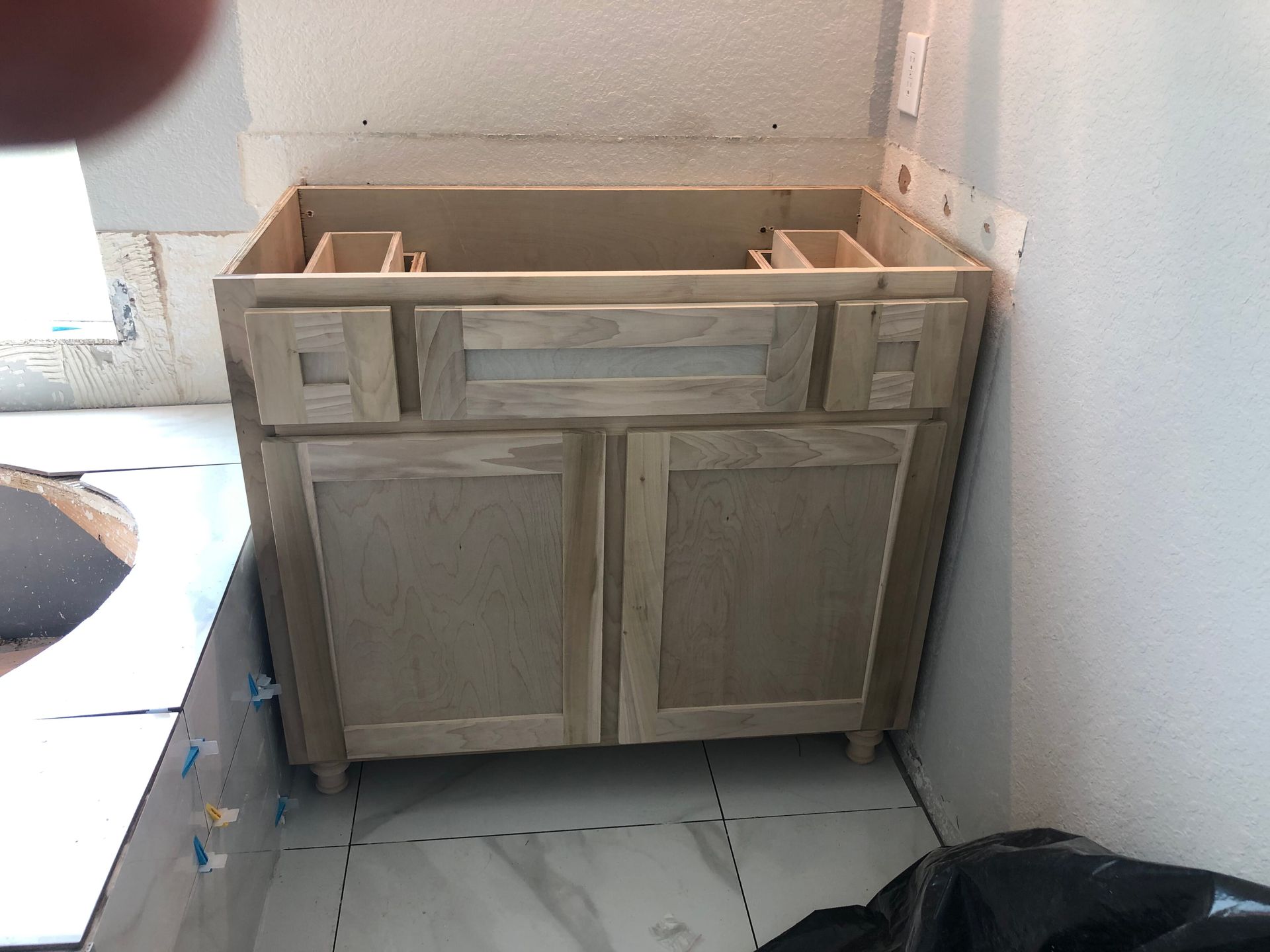 A wooden bathroom vanity cabinet installed on an unfinished tile floor in a room undergoing renovation.