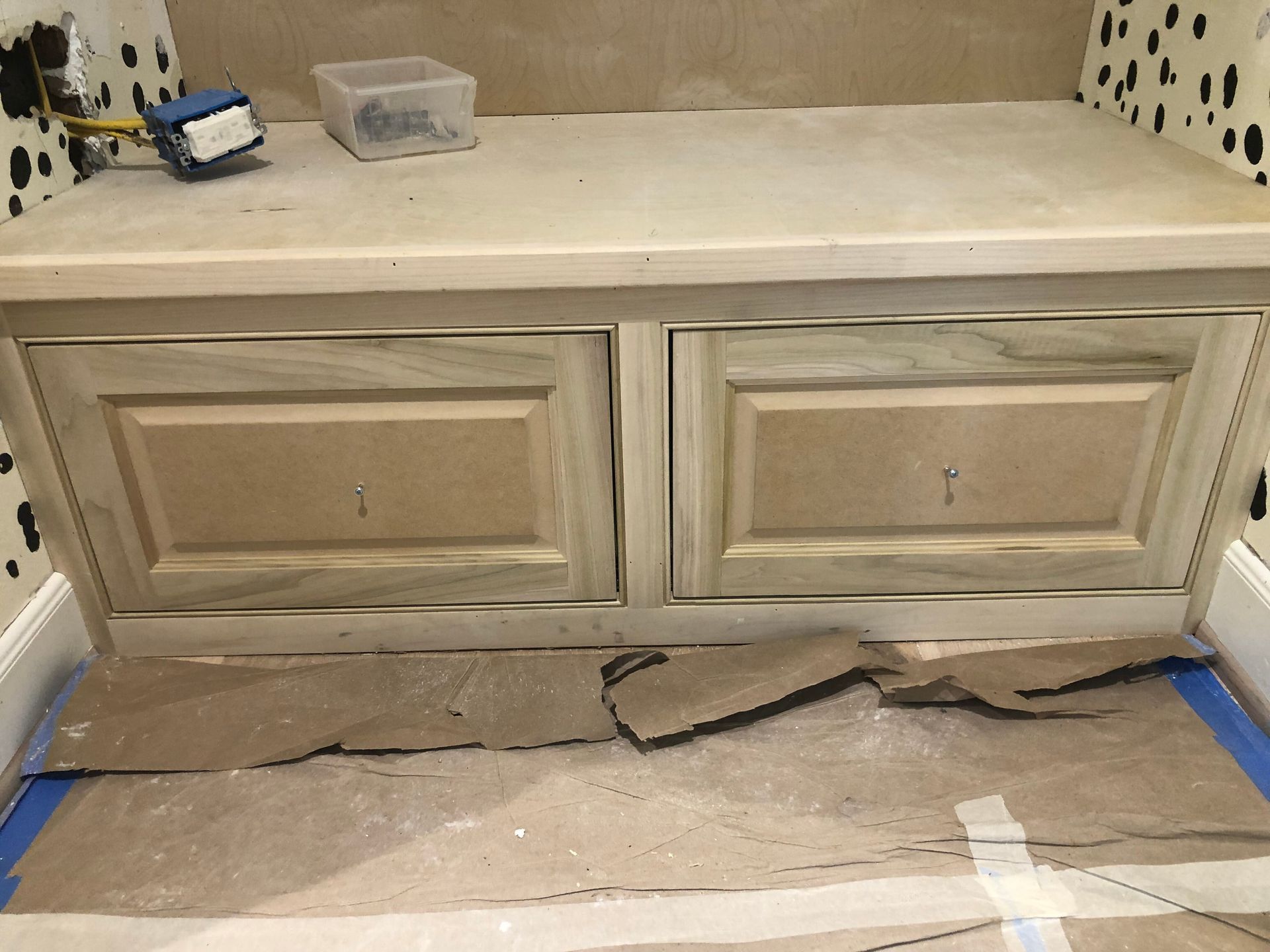 A wooden built-in bench with two cabinet doors under construction in a room with patterned walls and protective floor paper.