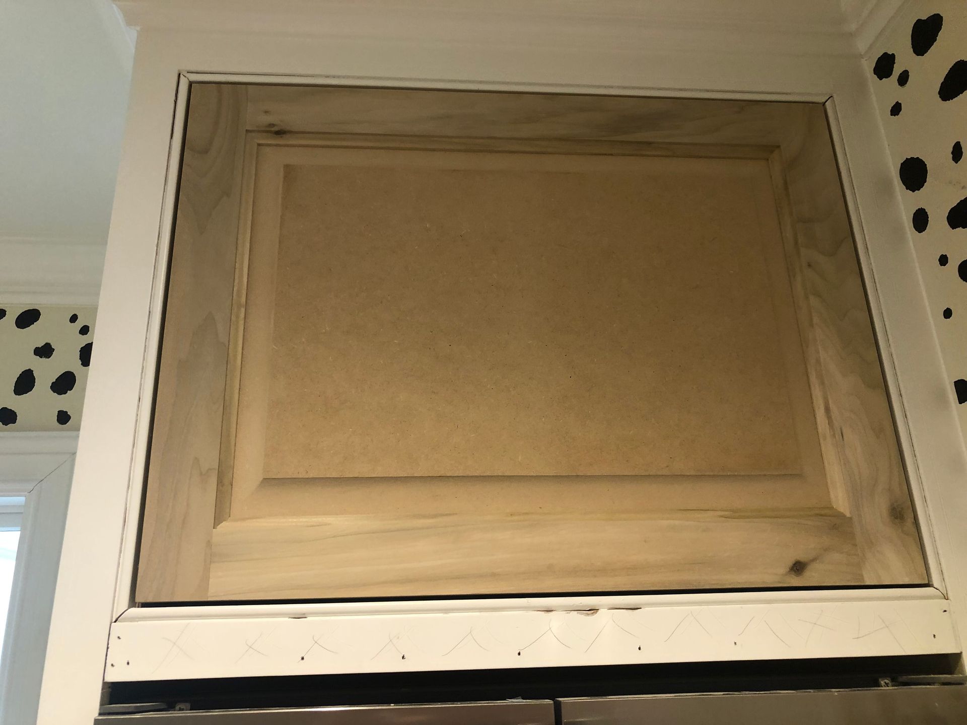 A wooden cabinet door above a refrigerator set against a white wall with black dalmatian-print wallpaper.