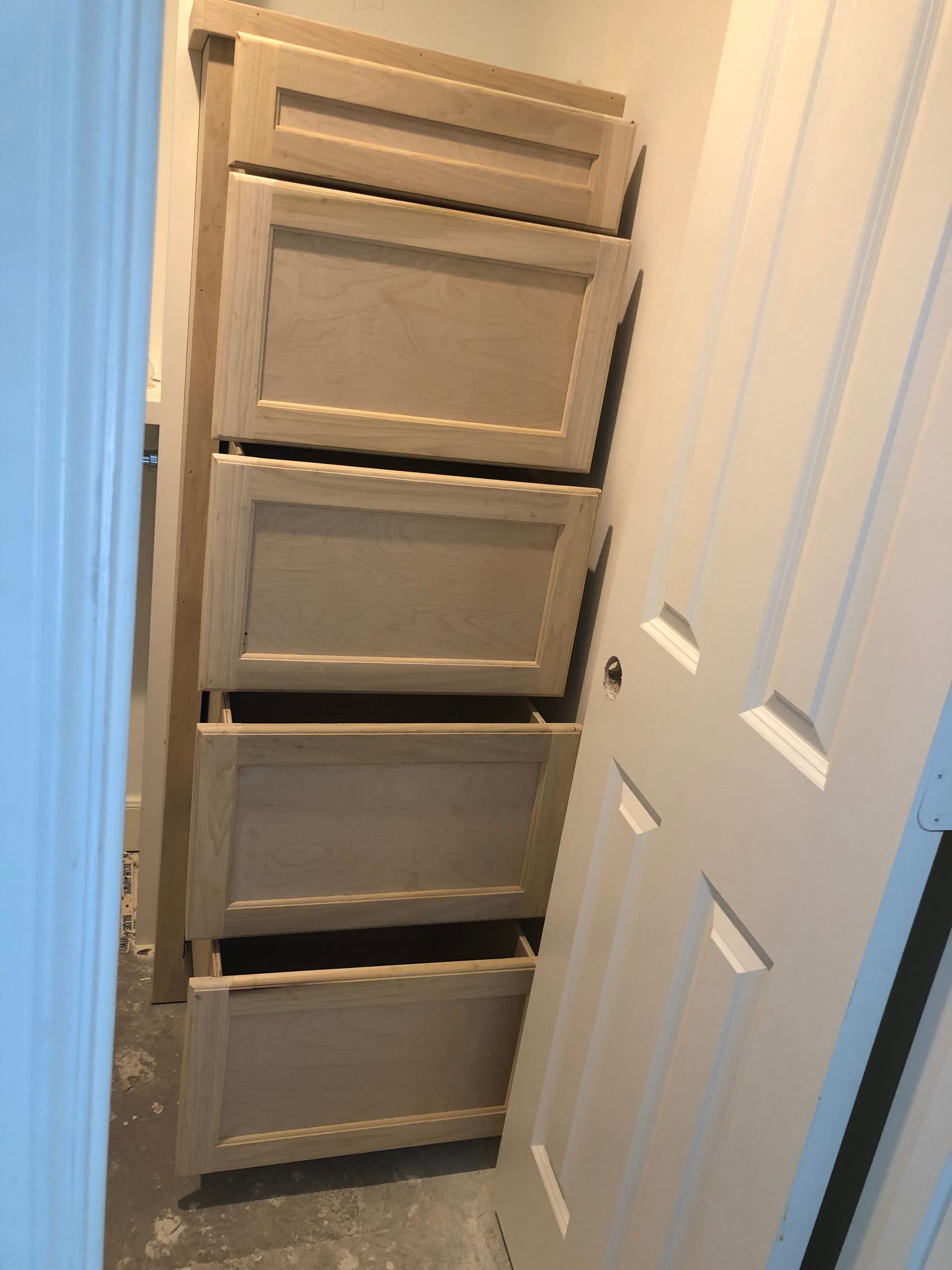 A tall, unfinished wooden drawer unit stands in a small closet space next to a white interior door.