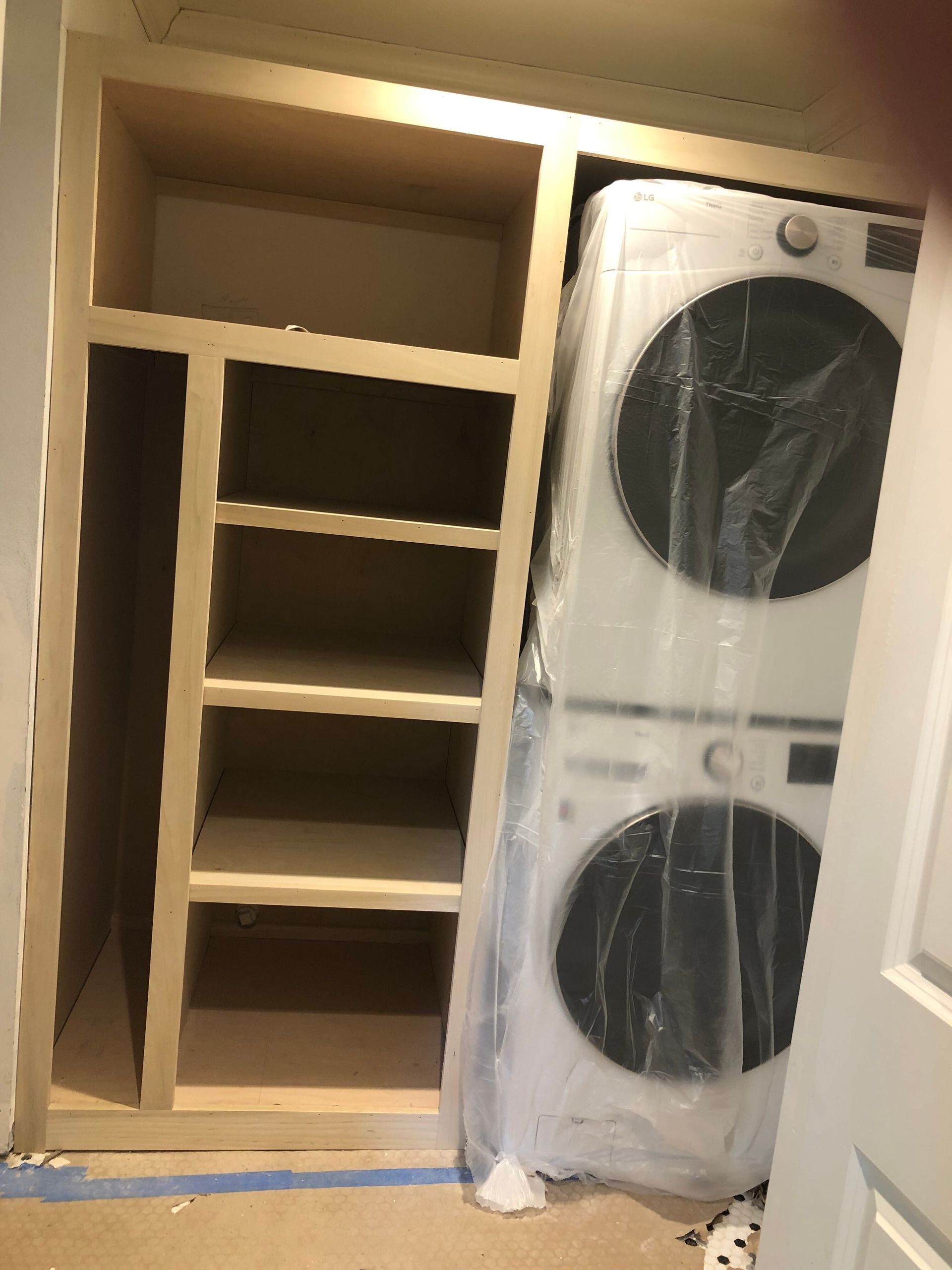A built-in wooden closet unit with empty storage shelves next to a stacked washer and dryer unit covered in plastic.