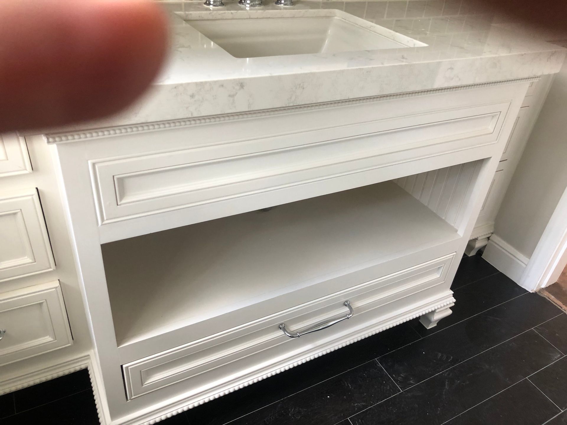 White bathroom vanity with marble countertop, open shelf, and drawer hardware against a black tiled floor.