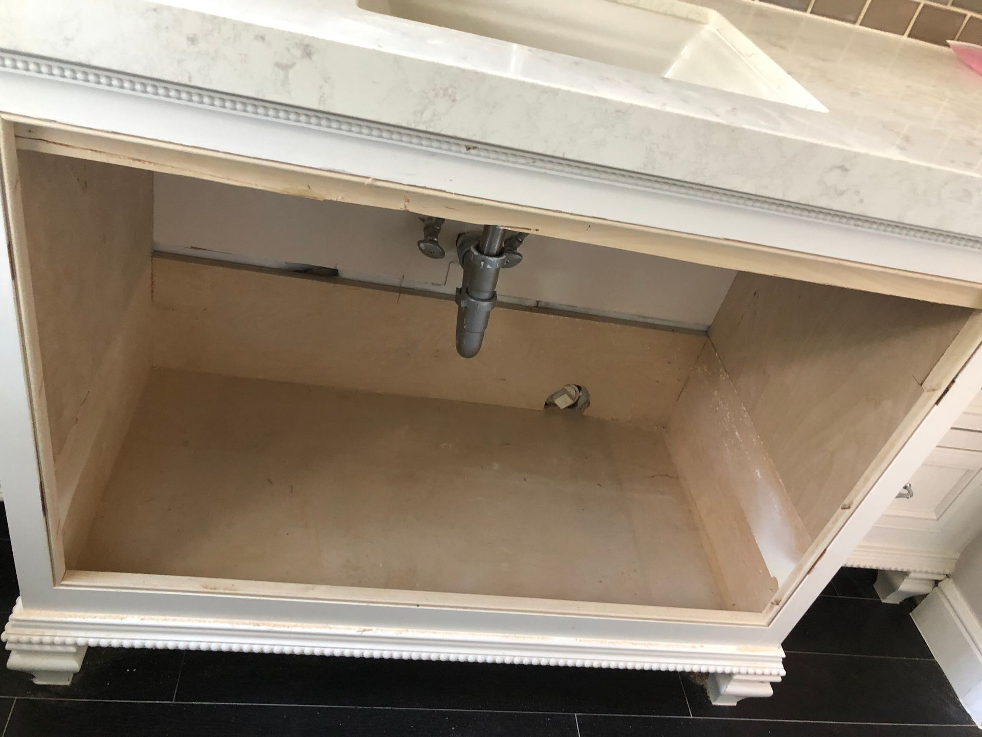 Open white bathroom vanity cabinet base with marble countertop and exposed plumbing against a tiled wall.