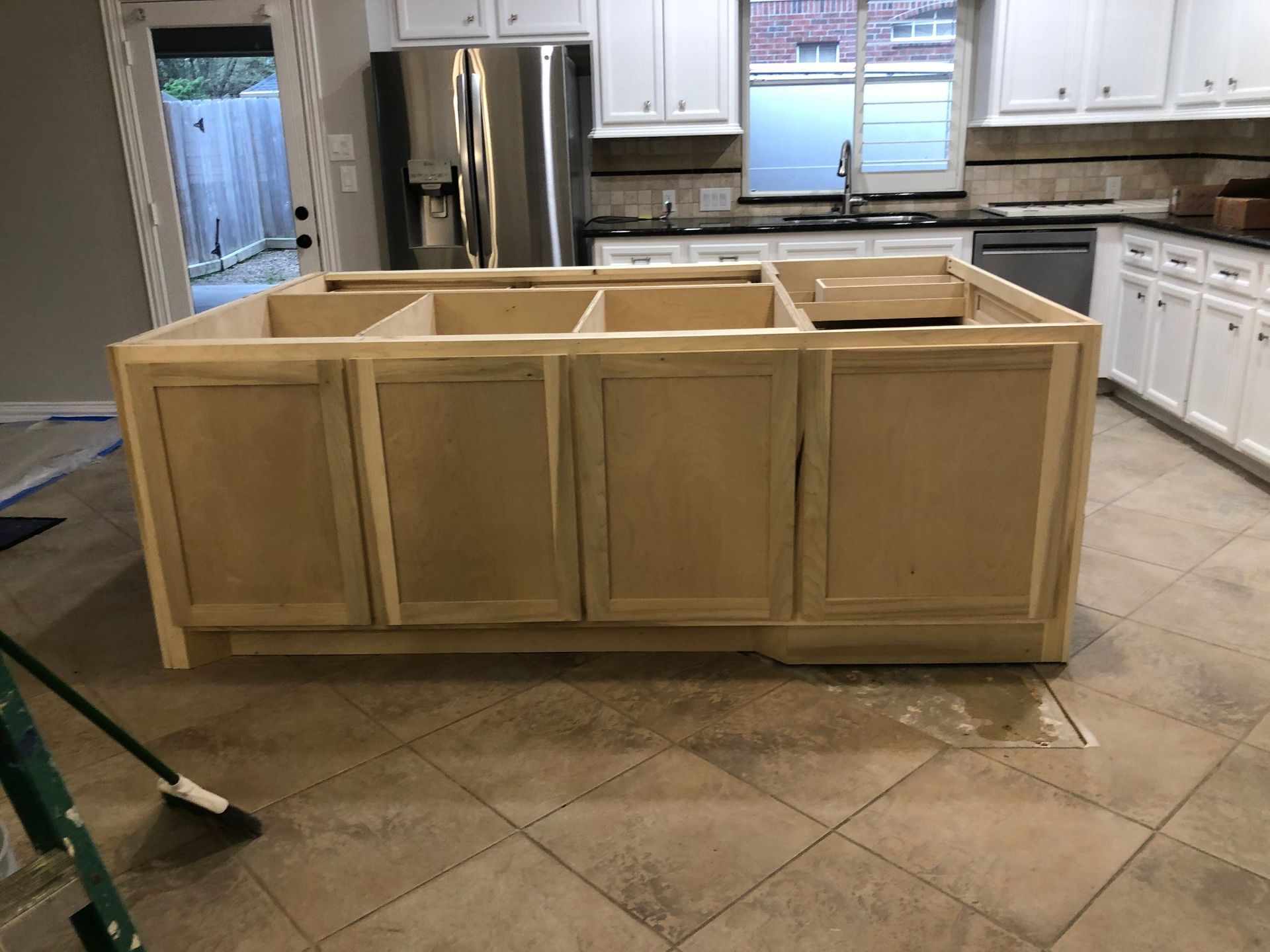 A large, unfinished wooden kitchen island base sits in the center of a kitchen with tile floors and white cabinets.