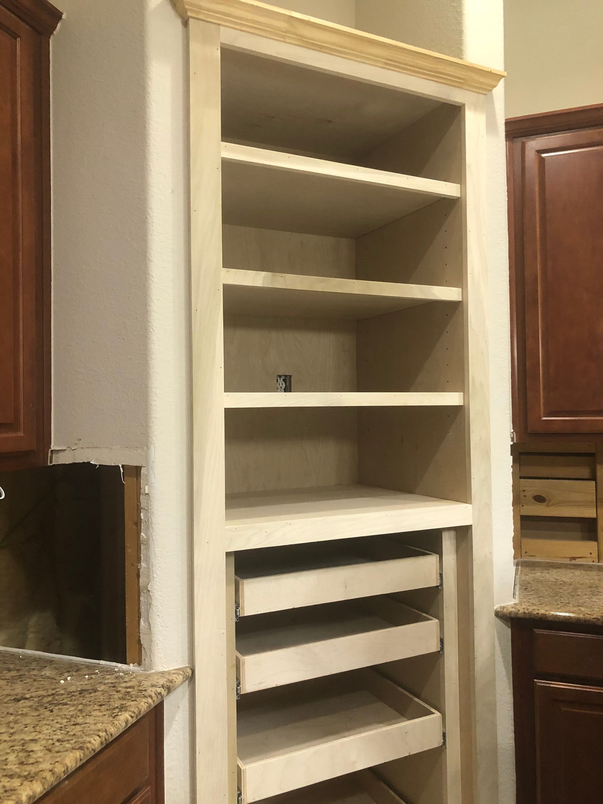 Unfinished wooden pantry cabinet with open shelving and pull-out drawers, flanked by dark brown kitchen cabinetry.