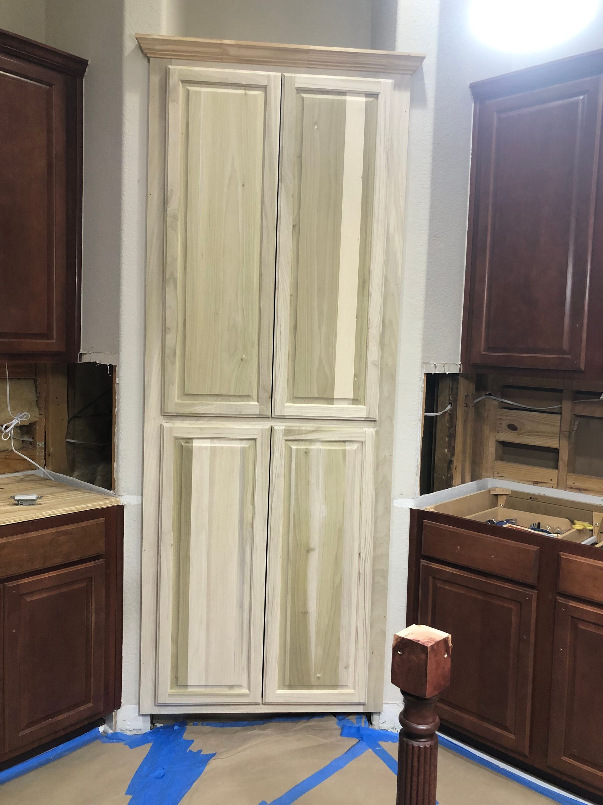 A light wood pantry cabinet stands between two dark wood lower cabinets in a kitchen under renovation.