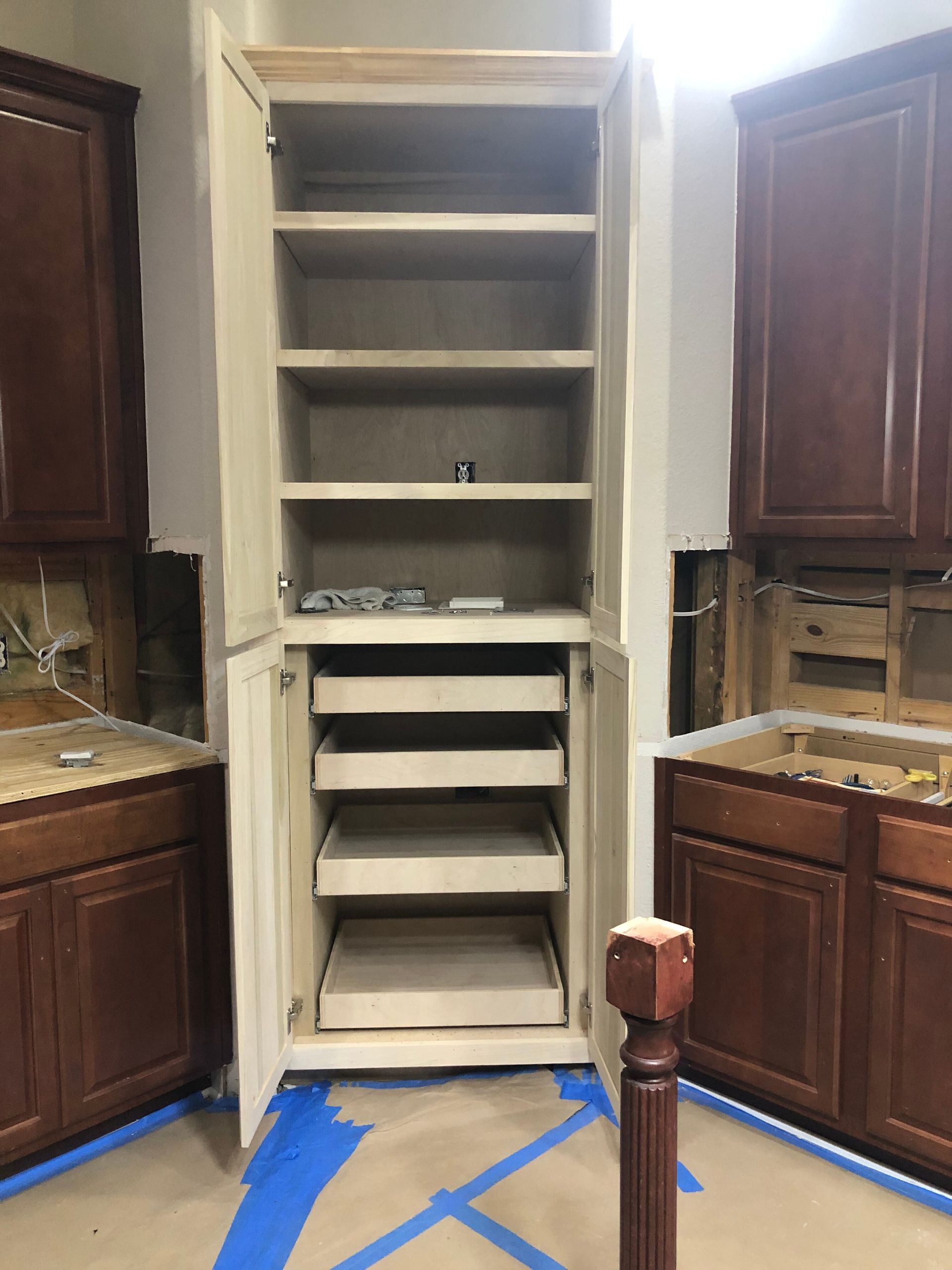 A tall, light-wood pantry cabinet with open shelves and sliding drawers sits between two dark-wood kitchen base cabinets.