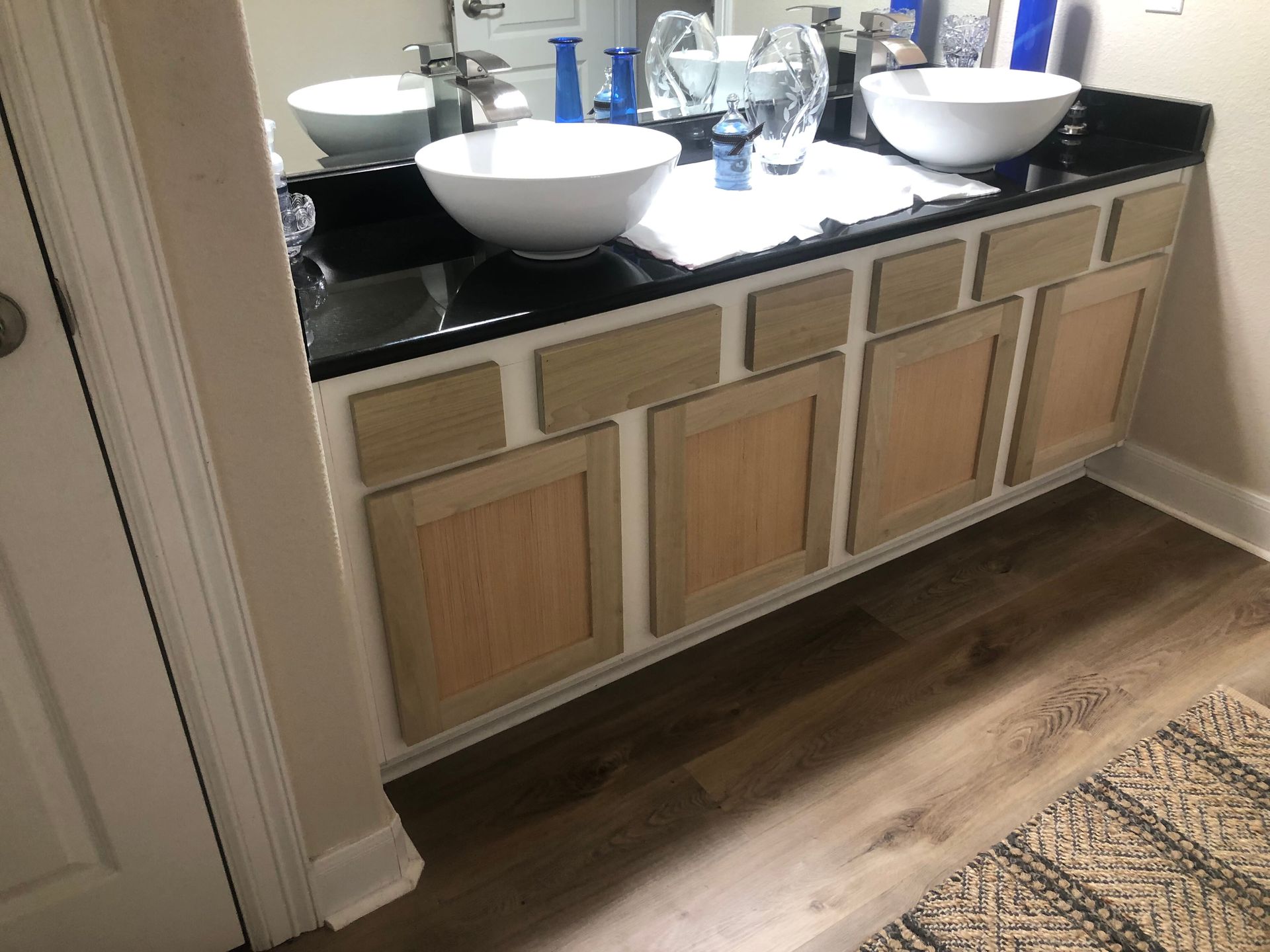 A bathroom vanity with a black countertop, two white vessel sinks, and light wood cabinet doors with a white frame.